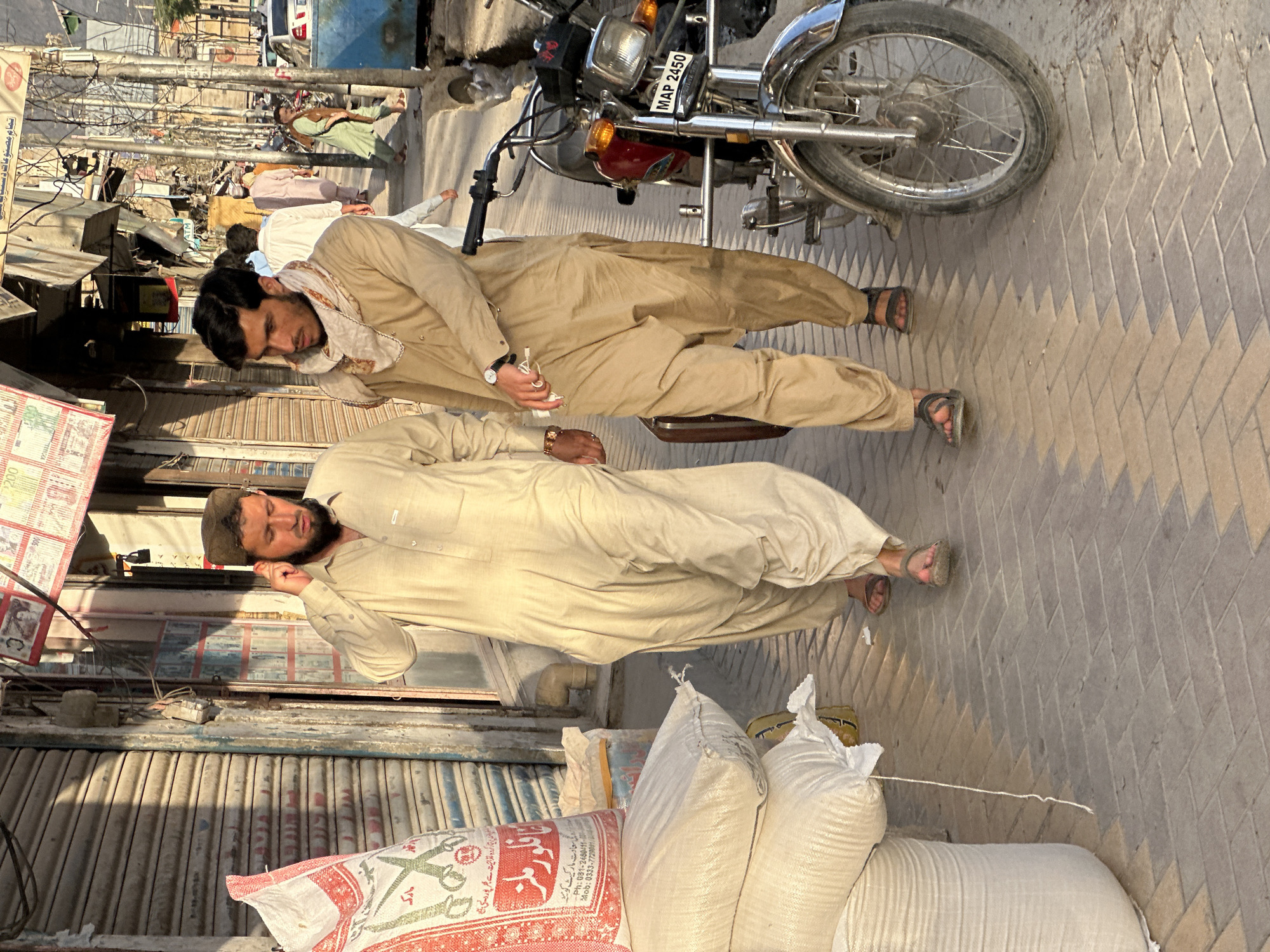 The image shows a street scene in Quetta City Tehsil, Pakistan. Two men wearing traditional Pakistani clothing are lying on the ground next to a motorcycle. One man is wearing a light brown shalwar kameez with a white patterned scarf, and the other is wearing a light cream-colored shalwar kameez. Next to them are several large white bags, possibly containing flour or grain, and there is a red and white bag with Pakistani writing on it. The motorcycle has a license plate that reads "MAP 2450." The street is paved with gray bricks and lined with storefronts with closed metal shutters. Power lines and other wires are visible overhead. The overall lighting suggests it is daytime.