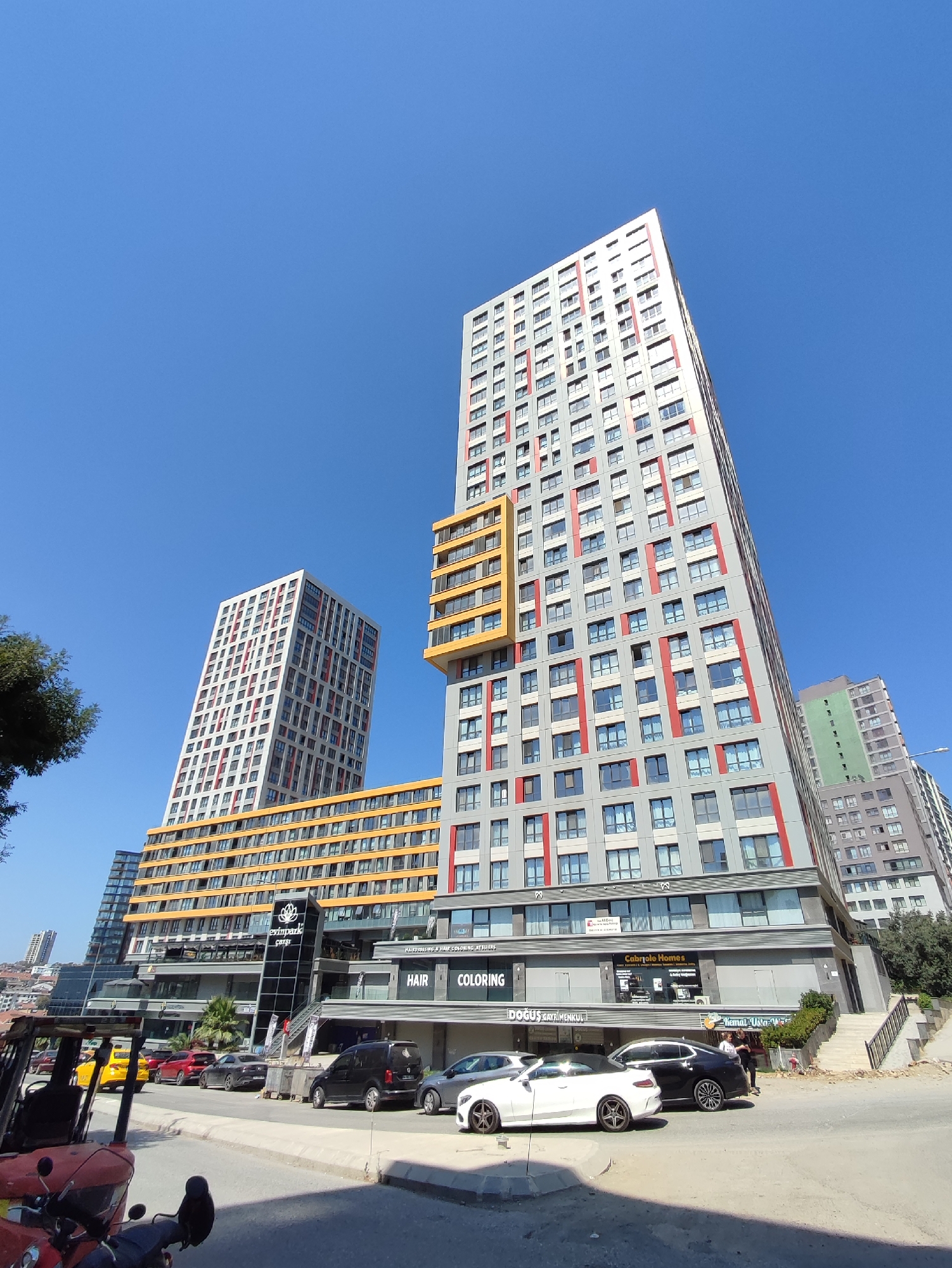 In Kadıköy, Turkey, a modern city skyline is dominated by several tall, multi-story buildings against a clear blue sky. The main structures are residential or mixed-use complexes featuring grey facades with striking red and yellow architectural accents. One prominent building has a yellow section near its top, while another features red vertical stripes. The lower levels of these buildings house commercial spaces, with signage visible including "Evinpark Çarşı," "Cabriolet Homes," "Doğuş Gayrimenkul," "HAIR," and "COLORING." A street runs in front of these buildings, with parked cars including a yellow taxi, a black minivan, a white convertible, and other passenger vehicles. A red three-wheeled vehicle, possibly a tuk-tuk or motorized cart, is parked on the left side of the street. A few individuals are visible near the entrance of one of the buildings, interacting with each other. Green trees and shrubs are present, bordering the street and near some of the buildings, suggesting an urban park or landscaped area. The overall scene depicts a contemporary urban environment with a mix of residential and commercial development.