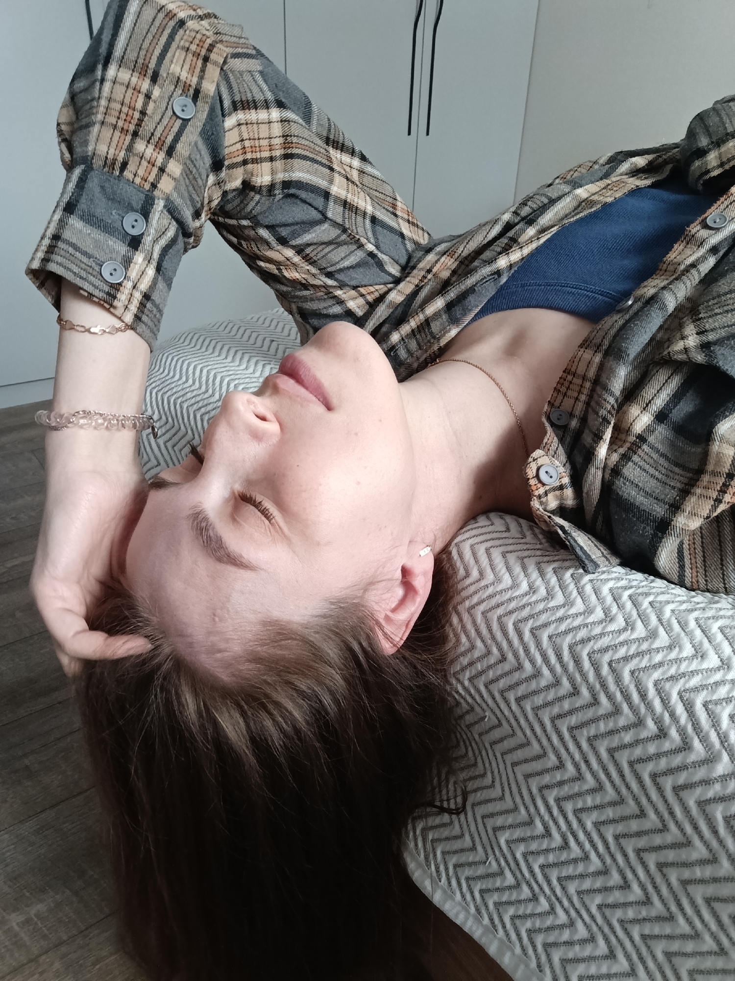A woman is lying on a cushion with a zigzag pattern, her head tilted back. She is wearing a blue top and a plaid shirt with buttons. Her right hand is touching her hair. She is wearing a gold bracelet on her left wrist and a delicate necklace. She has light skin, closed eyes, and her hair is brown. The location may be inside a building in the city of Chaikovsky, Russia, given the prompt. The scene includes a wooden floor and a white wall with black accents.