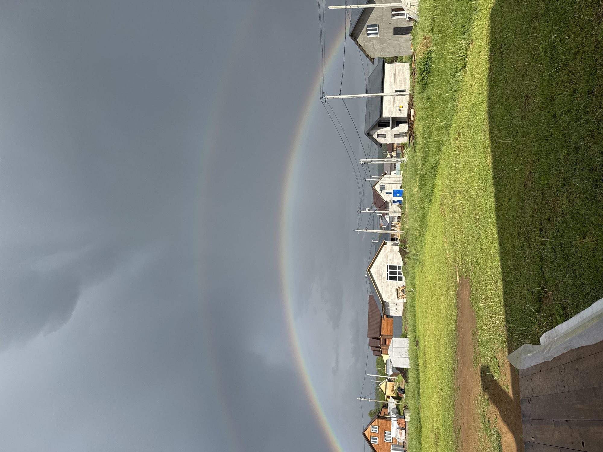The image shows a scene in the city of Ферма, Russia, where a double rainbow is visible in the gray sky. Below the rainbow, a row of houses is situated along a green grassy field. Power lines run along the side of the houses. The houses are in varying stages of construction, with some appearing to be newly built or still under construction. In the foreground, a section of wooden decking or platform is visible.