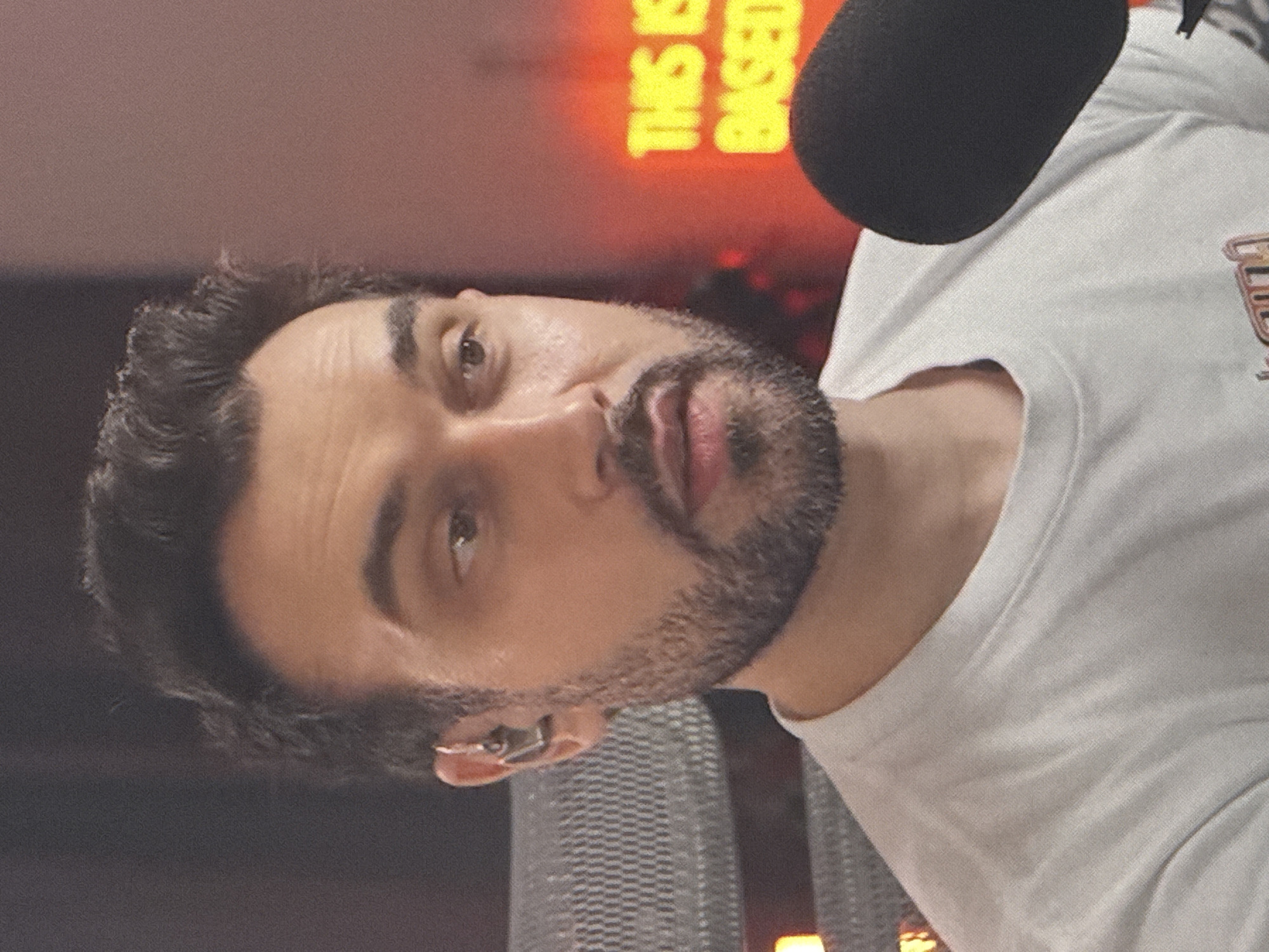 The image shows a medium shot of a man wearing a white t-shirt and an earbud. He has dark hair and a beard, and is looking slightly off-camera. In the background, a neon sign with the words "THIS IS BASED" is visible. The setting appears to be indoors, possibly a studio or recording space, with a microphone visible in the foreground on the right. There is no identifiable information in the image that would allow for the determination of its location in Saratov, Russia.