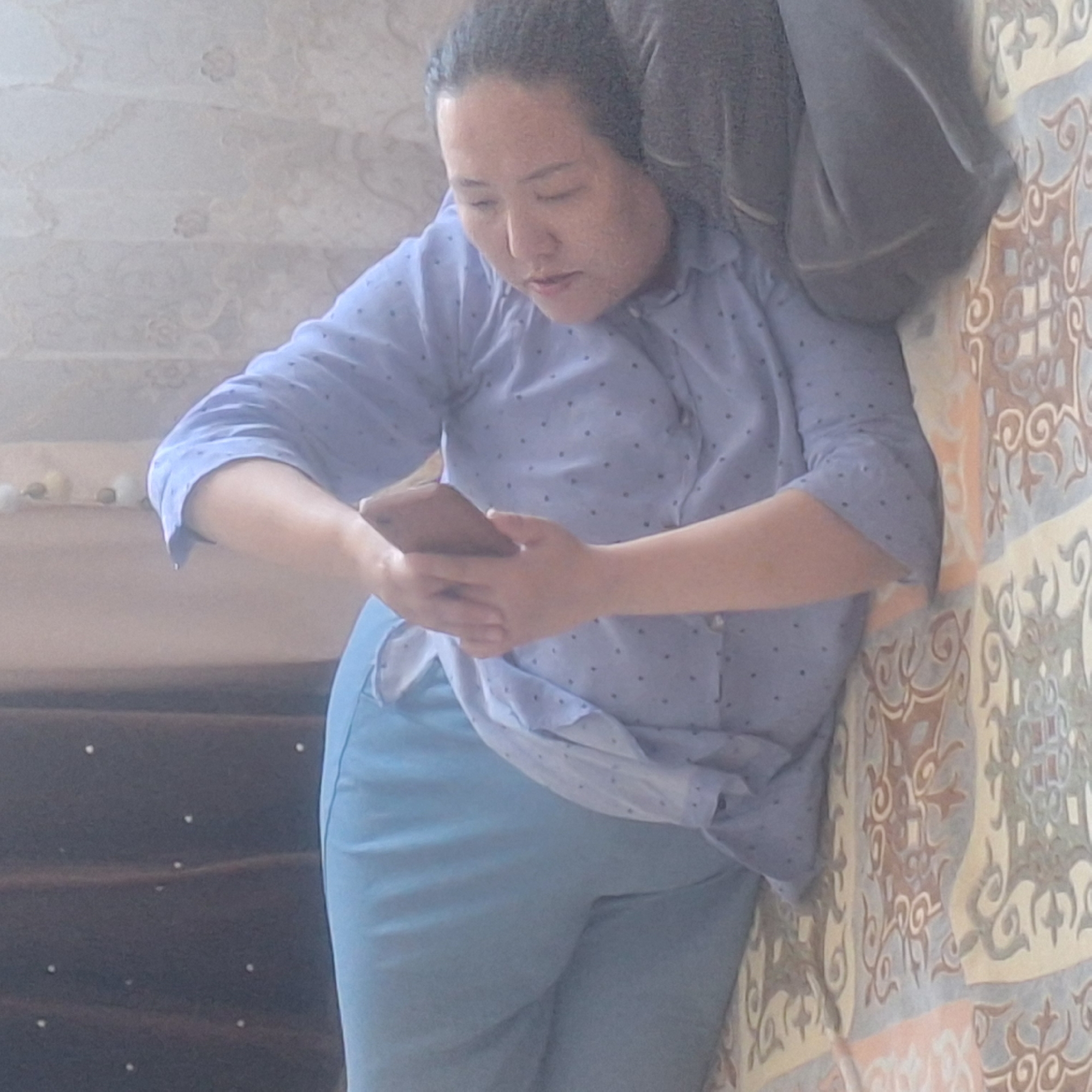 A person is lying on their side on a bed, holding a smartphone in their hands. The person is wearing a blue, polka-dotted shirt and light blue pants. The bed is covered with a patterned blanket. A dark grey pillow is under the person's head. In the background, there are brown curtains and a sheer, patterned curtain. The scene appears to be indoors.