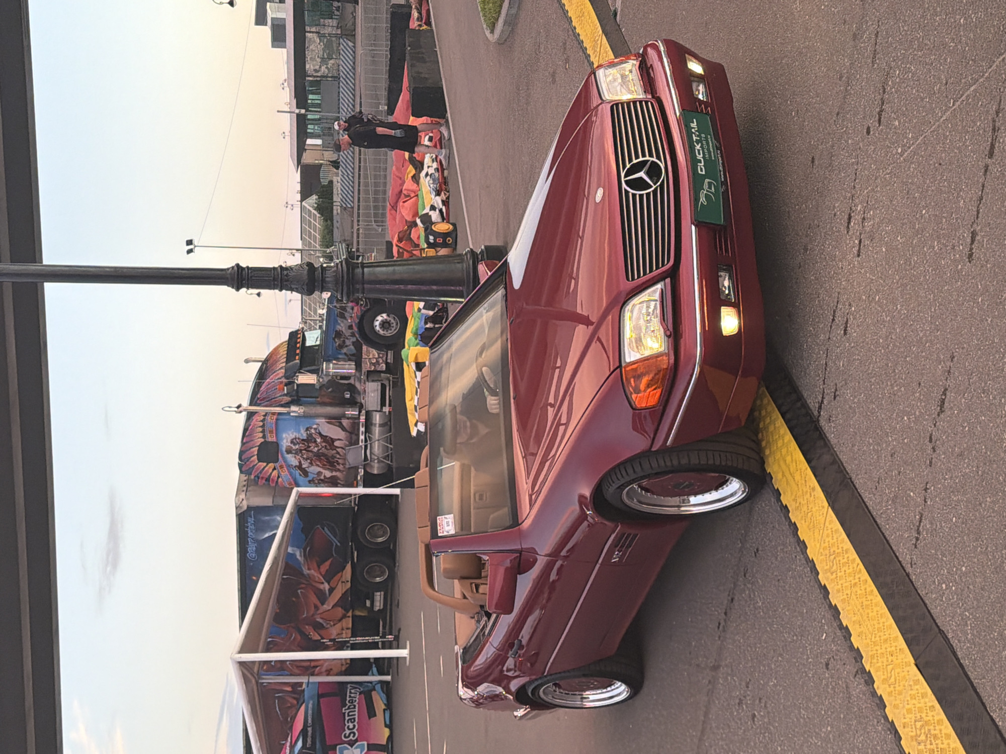 An exterior medium shot captures a dark red Mercedes-Benz SL convertible parked on an asphalt road. The car is angled from the front left, with its convertible top down, revealing tan leather interior. A driver, partially visible and wearing a dark shirt, is seated behind the steering wheel. The car has chrome rims with red accents and a license plate frame that reads "DUCK TAIL IMPORTS". In the background, a large truck with custom artwork of Native American figures and warriors is visible. To the left of the truck, a white tent structure with similar artwork is present. Further in the background, there are solar panels and some industrial buildings. Two men in casual attire, one wearing a hat, stand in the middle ground, near a black barrier. In the foreground, a yellow and black speed bump crosses the road. The sky is a pale blue with hints of orange and pink, suggesting a sunset or sunrise.