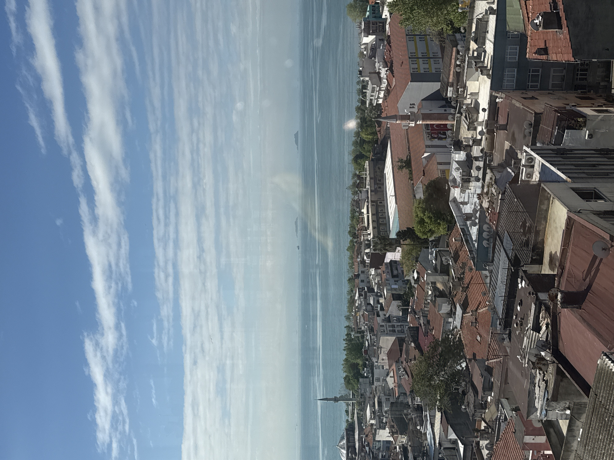 The image shows a cityscape with the sky on the left, a body of water in the center, and buildings on the right. The sky is blue with long, white clouds. The water appears calm and has a light blue hue. The buildings on the right are densely packed, with a mix of red-tiled roofs and various architectural styles. A minaret is visible, suggesting the presence of a mosque. Satellite dishes adorn many of the rooftops. The location appears to be an urban area near a coastline. The city is Unknown, Unknown.