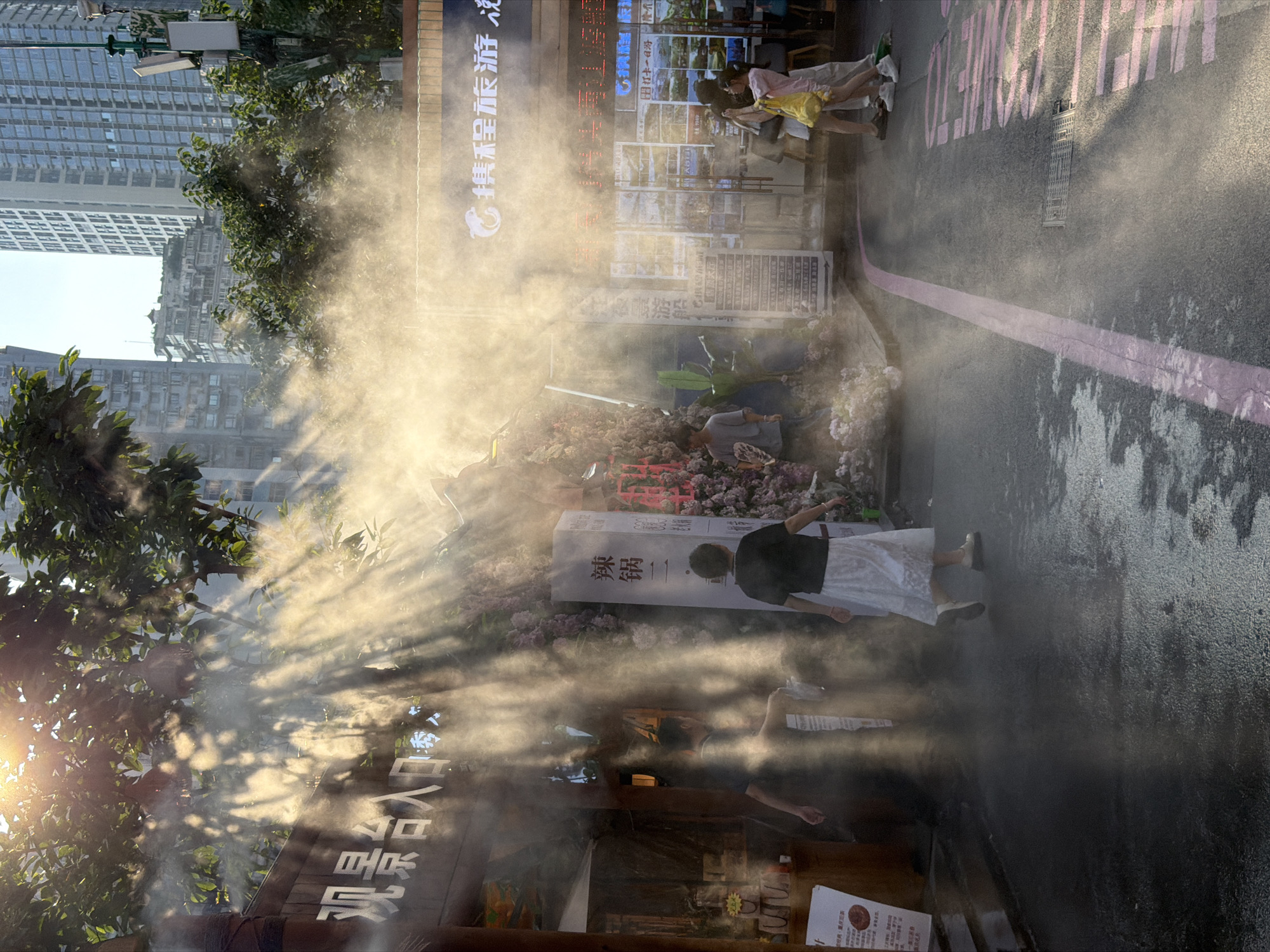 The image depicts a street scene in Yuzhong District, China, characterized by a bright sun casting strong rays through a misting system. Buildings, including modern high-rises and older structures, are visible in the background. In the foreground, several people are walking. One woman in a black shirt and white skirt is walking away from the camera, her figure illuminated by the sunbeams. Another person in a black shirt is walking past a stall with Chinese characters. To the right, two young women are walking together, one taking a photo with her phone, and another woman in a pink shirt is holding a yellow bag. The street is paved with asphalt and has a pink line painted on it. There are also plants, decorative flowers, and signs with Chinese text visible, suggesting a commercial or tourist area. The mist creates a hazy atmosphere, with the sunbeams creating dramatic shafts of light.