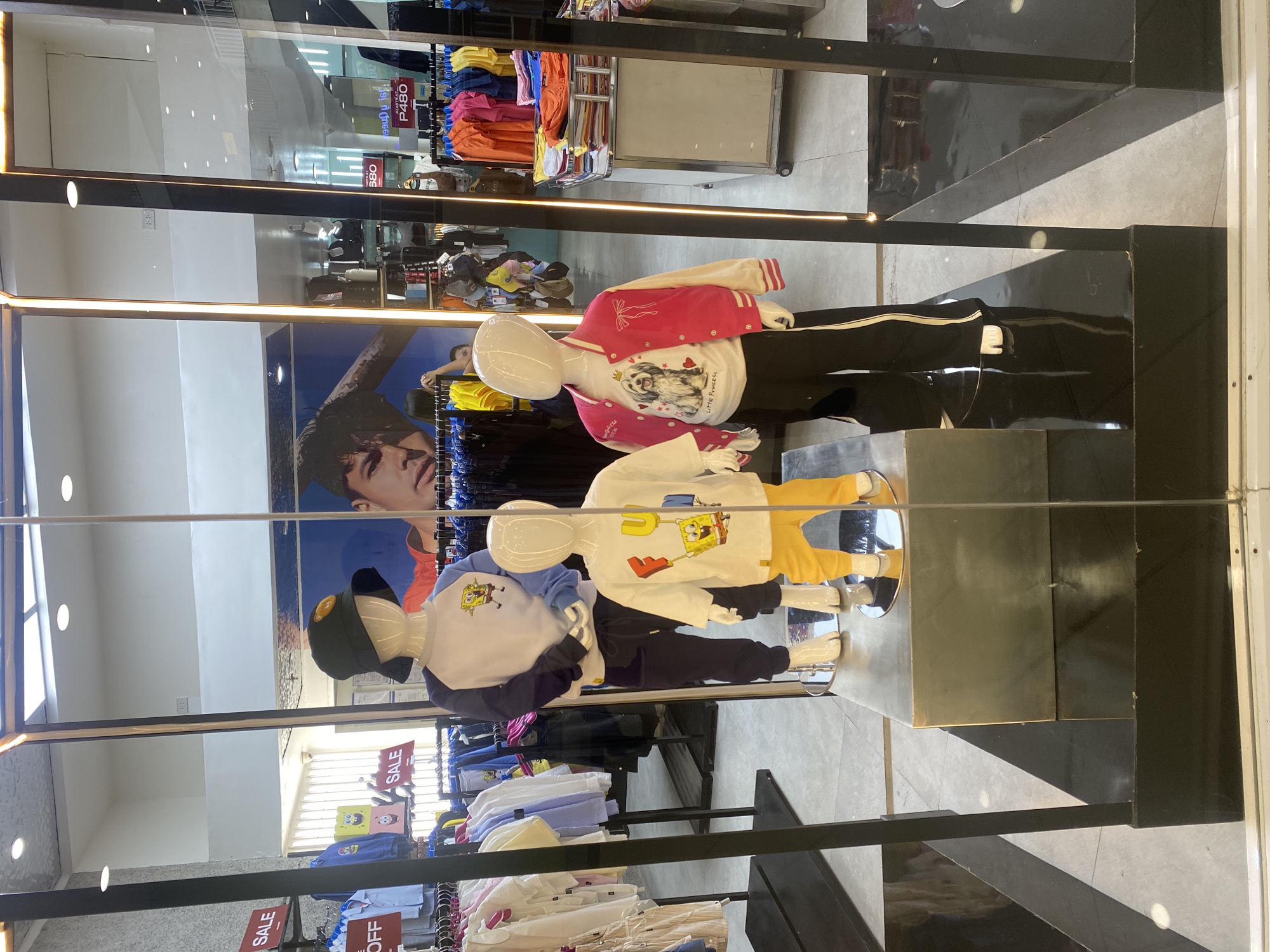 The image displays a retail clothing store interior, likely in a mall in Davao City, Philippines, presented through a glass display window. The entire image is rotated 90 degrees counter-clockwise, showing the store's floor on the right and the ceiling on the left.
Three child-sized mannequins are arranged horizontally within the display, showcasing children's apparel. The leftmost mannequin wears a white long-sleeved shirt with a graphic of a dog and the text "Little Princess" surrounded by red hearts, topped with a pink and white varsity-style jacket. It is paired with black pants featuring a white side stripe. The central mannequin sports a white long-sleeved shirt with a Spongebob Squarepants graphic and bright yellow pants. The rightmost mannequin is dressed in a white and dark blue long-sleeved baseball-style shirt, also with a Spongebob Squarepants graphic, dark blue or black pants, and a dark cap with a yellow button.
In the background, a large poster features a close-up of a person's face. Numerous clothing racks are visible, laden with colorful garments in shades of pink, orange, yellow, blue, and white, both directly and reflected in the glass. The mannequins stand on glossy black platforms. The store has a light grey tiled floor and a ceiling with recessed lighting.
Visible text includes multiple red "SALE" signs strategically placed on clothing racks throughout the store, a partially visible "OFF" on a red sign, and a price tag showing "P480," indicating Philippine currency. The scene depicts a static commercial display designed to attract customers to the children's clothing section.