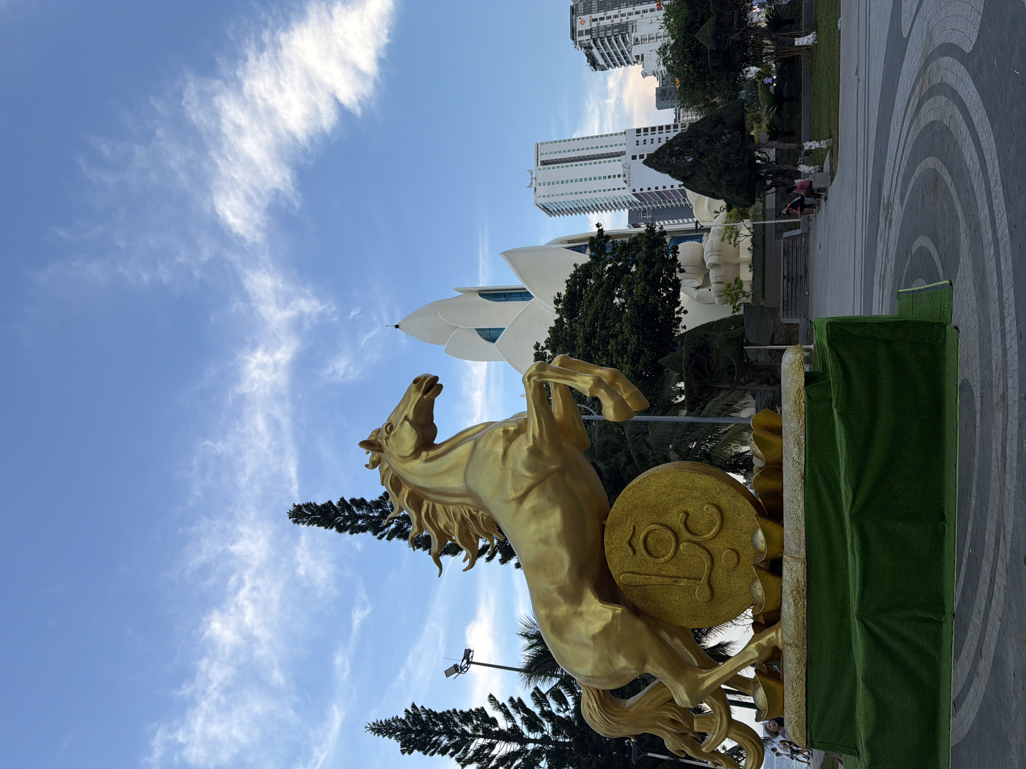 An outdoor, eye-level, wide-angle shot captures a golden statue of a horse rearing up on a large golden coin. The statue is positioned in the foreground, with its base covered in green artificial turf. Behind the statue, a distinctive white building with a petal-like roof design dominates the mid-ground. To the right of this building, several modern high-rise buildings are visible. In the background, a clear blue sky with scattered white clouds stretches overhead. Several dark green, conical trees are scattered throughout the scene, with palm trees visible behind the statue on the right. A paved walkway with a circular pattern is in the lower right quadrant of the image, with several people sitting on benches and walking in the distance. The location is identified as Nha Trang Ward, Vietnam.
