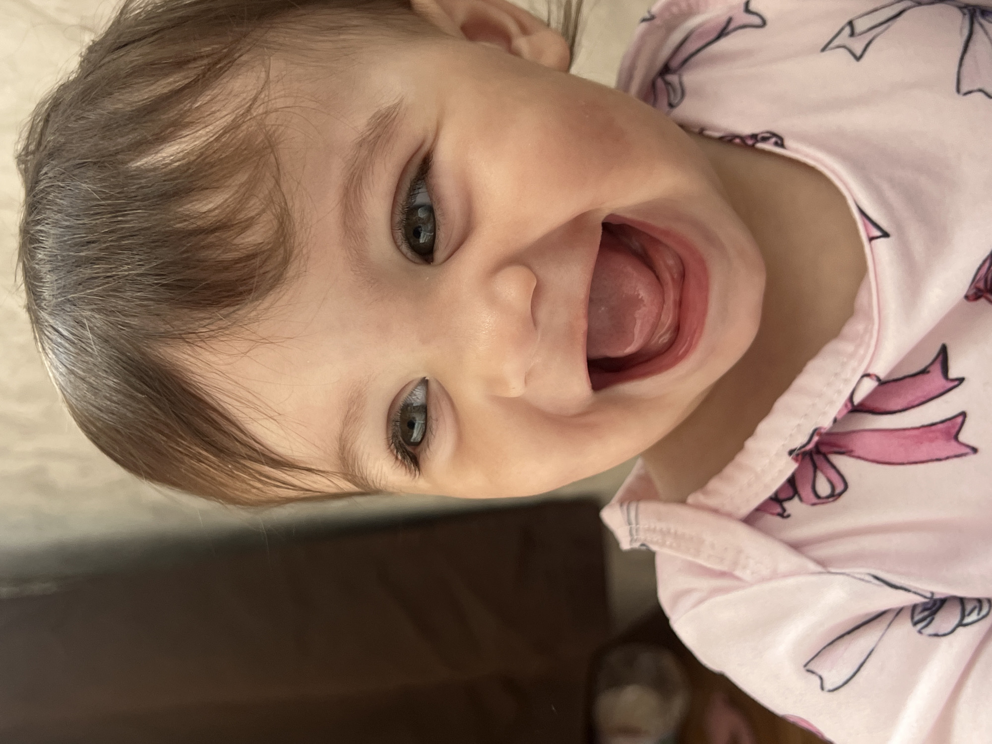 A close-up, tilted, indoor shot depicts a smiling baby girl with brown hair and grey-green eyes. Her mouth is open, revealing her tongue. She is wearing a pink pajama top with a ribbon pattern. A dark brown surface is visible in the lower portion of the frame. The image does not reveal any information about the location of Elena, Bulgaria.