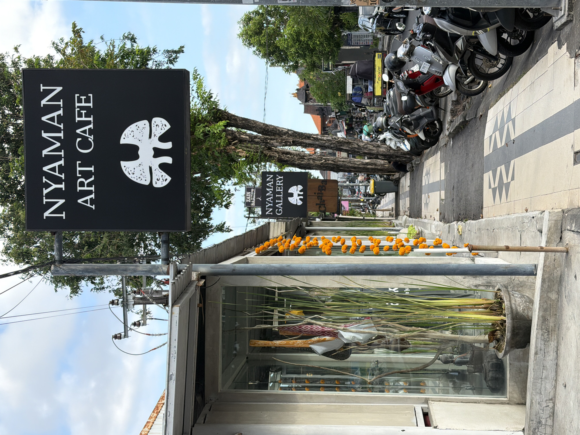 The image shows a street scene in Kuta, Indonesia, featuring the "Nyaman Art Cafe" and "Nyaman Gallery." The "Nyaman Art Cafe" sign, a black rectangular board with white lettering and a distinctive logo, is prominently displayed on the left. Further down the street is another sign that reads, "Nyaman Gallery." Orange garlands adorn the front of a building with large glass windows. Inside the windows, plants and fabrics are displayed. Parked motorbikes line the right side of the street, along a sidewalk with a black and white triangular pattern. Trees are interspersed along the road. The sky is partly cloudy.