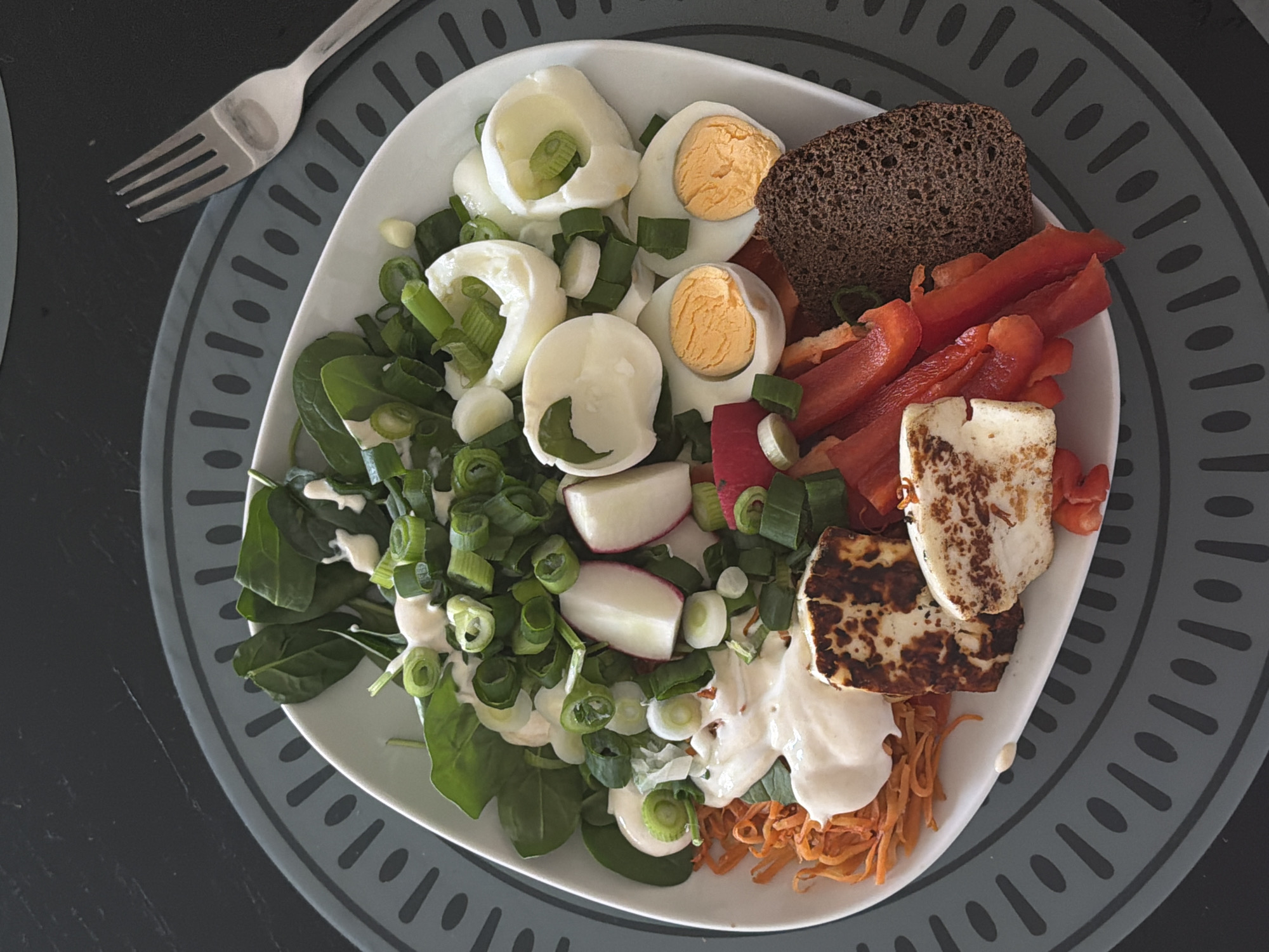 A top-down shot captures a white bowl filled with a salad on a grey circular placemat. The salad comprises spinach leaves, sliced green onions, halved hard-boiled eggs, radish slices, red bell pepper strips, shredded carrots, two grilled halloumi cheese pieces, a dollop of white sauce, and a slice of dark bread. A silver fork rests on the placemat beside the bowl. The background is a dark surface. The location is not evident in the image.