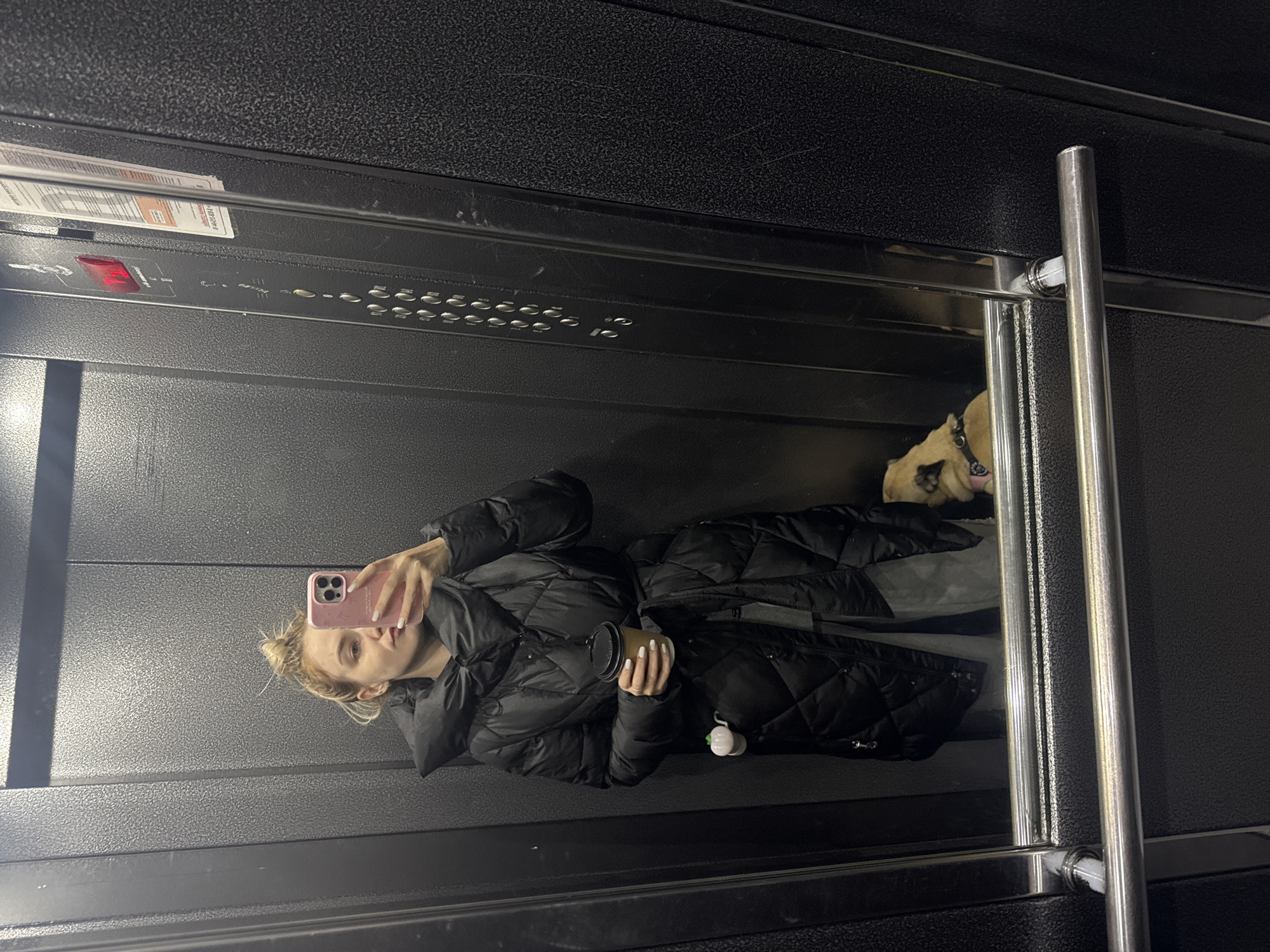 A photograph depicts a woman taking a selfie in what appears to be an elevator. She is wearing a black puffer jacket and holding a light-colored coffee cup in her left hand and a pink phone in her right. The woman has blonde hair pulled up into a messy bun. Behind her and to the right is a fawn-colored dog wearing a collar. The interior of the elevator is primarily dark, with horizontal bands of textured metal. Control panels are visible above and beside her. Metal handrails are on the right. The location is unknown and not discernible from the image.