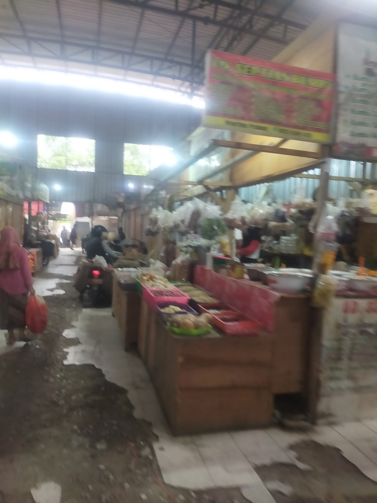 The image depicts an indoor marketplace, likely a traditional market, in Trayeman, Indonesia. The scene is bustling with activity, though the image quality is somewhat blurred. In the foreground, a person wearing a pink headscarf and skirt walks away from the camera, carrying a red plastic bag. To the right, market stalls are visible, displaying various goods. One stall features a wooden counter with trays of what appear to be snacks or food items. Another stall has rows of packaged goods hanging, possibly snacks or spices. The stalls are set up under a metal roof supported by wooden beams. Natural light streams in from openings in the roof or walls, creating bright areas. Several motorbikes are parked within the market area. In the background, other people can be seen walking through the market, and some are near stalls. Signs are visible, one with red and yellow lettering, suggesting shop names or advertisements. The floor of the market appears to be tiled in some areas and dirt or rough ground in others, with some patches of water or dampness. The overall impression is of a vibrant, active marketplace.