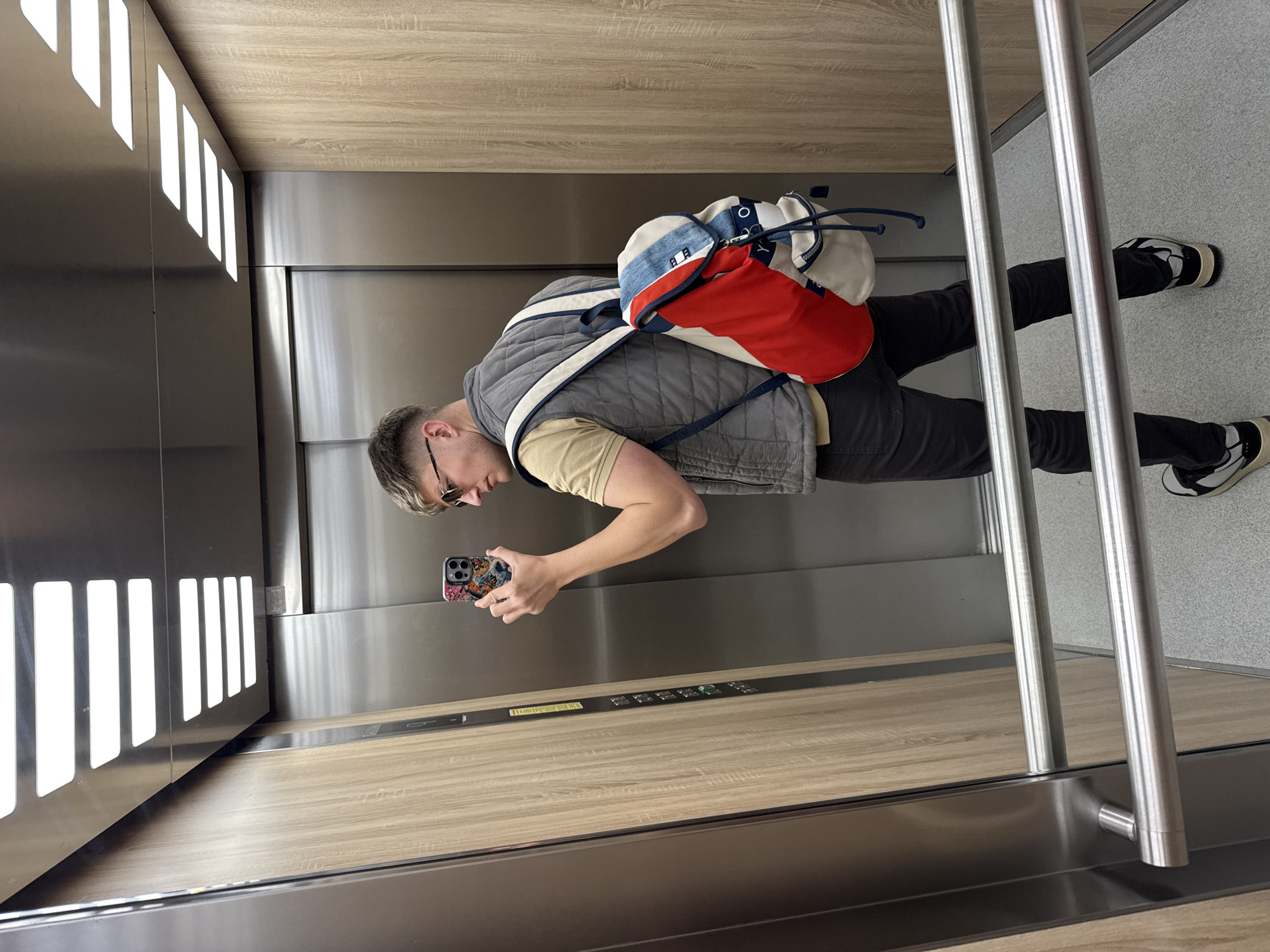 The image shows a person, presumably male, taking a selfie in an elevator. He's wearing a grey quilted vest over a tan shirt, dark pants, and black-and-white sneakers. He has a colorful backpack. The elevator has a modern design with stainless steel walls, wood paneling, and vertical light strips on one side. Two metal railings are visible on the right side of the image. The background outside the elevator appears to be a light grey or white surface. The location in the city of Plovdiv, Bulgaria is not visually detectable in the given image.