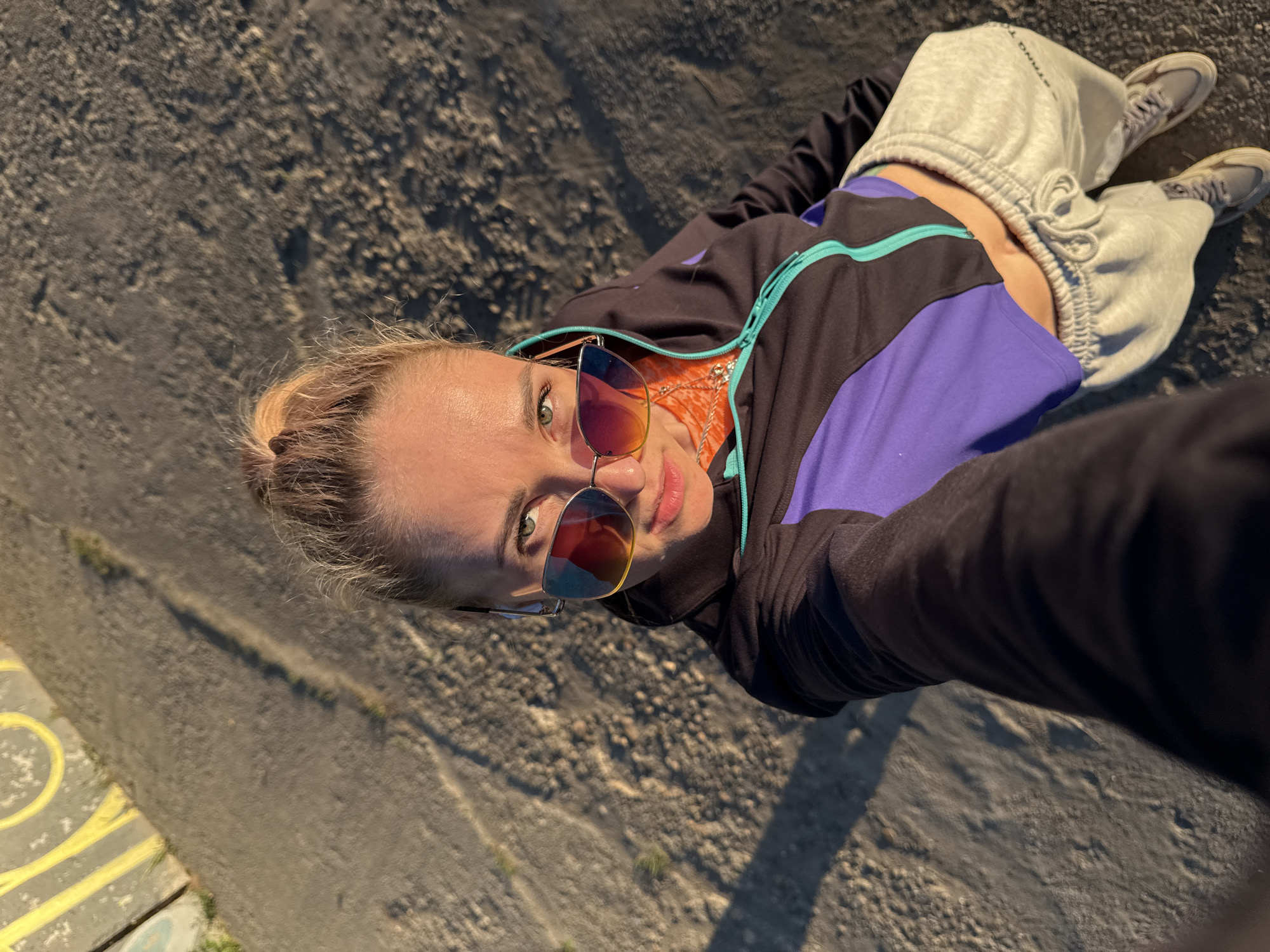 A young woman is depicted in a high-angle selfie, looking up directly into the camera while lying or sitting on a rough, textured ground surface. She has fair skin, light-colored hair tied back, and green eyes, partially concealed by large, reflective sunglasses with colorful, iridescent lenses. Her attire includes a black and purple athletic jacket with a bright turquoise zipper and trim, an orange top underneath, light grey sweatpants, and light grey sneakers. The sweatpants display text, with "I STAND FOR" visible on the left leg.
The setting appears to be outdoors, with warm, directional sunlight suggesting late afternoon or golden hour, casting shadows and highlighting the uneven ground. The background is composed of dark, granular material, possibly asphalt or gravel. Subtle yellow markings are visible on a paved section in the lower left corner, indicating a road, pathway, or a painted area. The overall mood is relaxed and casual. The image provides a glimpse of an outdoor moment in Chaikovsky, Russia.