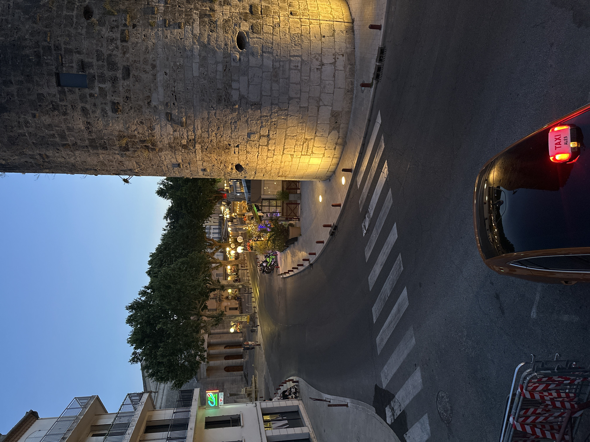 The image captures a street scene in the city of Anduze, France, at dusk. A large, ancient stone tower dominates the right side of the frame, illuminated by warm, yellow lights at its base. To the left, a multi-story building with balconies and a green bank sign is visible. A pedestrian crossing with white stripes is painted on the dark asphalt road, which curves through the scene. Several trees with lush green foliage are planted along the street, with lampposts casting a glow on the pavement. In the foreground, the roof of a brown taxi with a lit "TAXI ALES" sign is visible. A metal barricade with red and white striped tape is positioned on the left side of the road. A few motorcycles are parked near a row of outdoor seating and potted plants in the middle ground. Two individuals are walking in the distance on the left side of the street, near a building with columns. The sky above is a deep blue, indicating twilight.