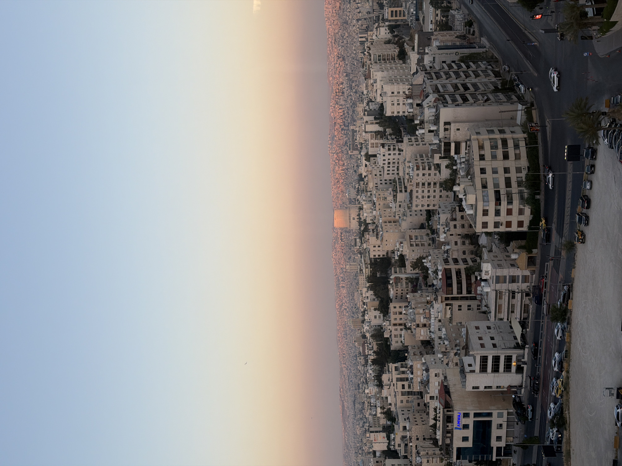 A high-angle, eye-level shot captures an urban cityscape at dusk. The sky transitions from a pale blue at the top to a soft gradient of pink and orange near the horizon, indicating the setting sun. A sprawling city with dense residential and commercial buildings stretches across the landscape, with countless structures dotting rolling hills in the distance. The buildings are predominantly made of stone and concrete, with varying architectural styles and heights. Some buildings feature balconies and windows that are visible. A paved road with multiple lanes is present in the foreground, with a few cars driving on it. There are also parked cars in a lot adjacent to the road. Trees and other greenery are interspersed throughout the city, adding a touch of nature to the urban environment. The overall impression is one of a large, active city bathed in the warm glow of twilight.