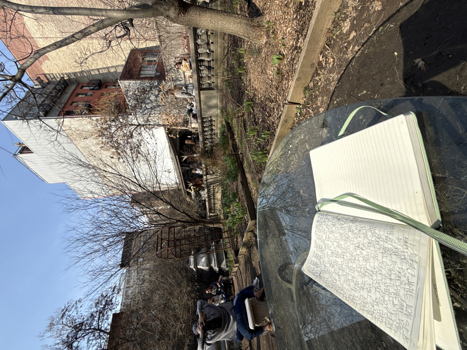 The image depicts an urban community garden or park in New York City, United States of America, under clear daylight, indicating late winter or early spring.
In the immediate foreground, an open journal with a light green cover and a matching ribbon bookmark rests on a dark, reflective table surface. The left page of the journal is filled with dense, repetitive handwriting, primarily reading "IT'S NOT THAT DEEP," while the right page is blank and lined.
The mid-ground reveals a garden with several raised wooden beds containing emerging green shoots. Bare deciduous trees with intricate branch patterns are scattered throughout. To the left, an old, rusted metal sculptural element and two weathered stone pedestals are visible. A white stone balustrade with ornate spindles runs horizontally, separating a lower garden area from an elevated section.
Multiple individuals are present. On a white stone bench in the left mid-ground, a person in a grey hooded sweatshirt and blue jeans sits with arms clasped behind their head, facing away from the camera. Two other individuals are partially visible, seated further to the left on the same bench. Behind the balustrade, several more people are gathered: one individual sits on the balustrade itself, another in a light brown jacket and sunglasses is seated to the right, and additional figures are congregated under a rustic wooden shelter with a sloped roof.
The background is composed of multi-story urban buildings characteristic of New York City, featuring brick facades, some with lighter siding, and visible fire escapes. Strong sunlight casts defined shadows across the scene.