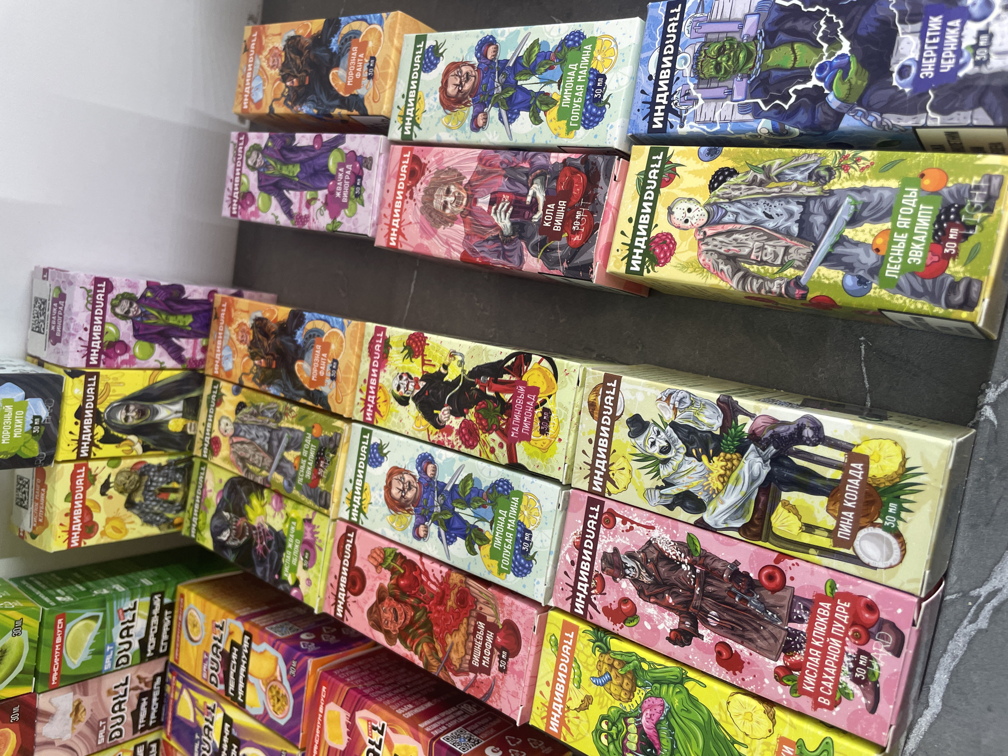 The image shows a collection of boxed items stacked on shelves, likely in a retail setting in Perm, Russia, given the Cyrillic writing on the boxes. The boxes appear to contain flavored vaping liquids from the brand “ИНДИВИDUALL” and “DUALL.” The boxes feature cartoonish, horror-themed characters such as Frankenstein, Jason Voorhees, and Freddy Krueger. The flavors are indicated by Cyrillic text, including flavors like blueberry energy drink, forest berries eucalyptus, pineapple coconut, and various fruit combinations. The boxes also display volume information, typically "30 мл" (30 ml). The shelves appear to be within a well-lit indoor space, potentially a store or distribution center.