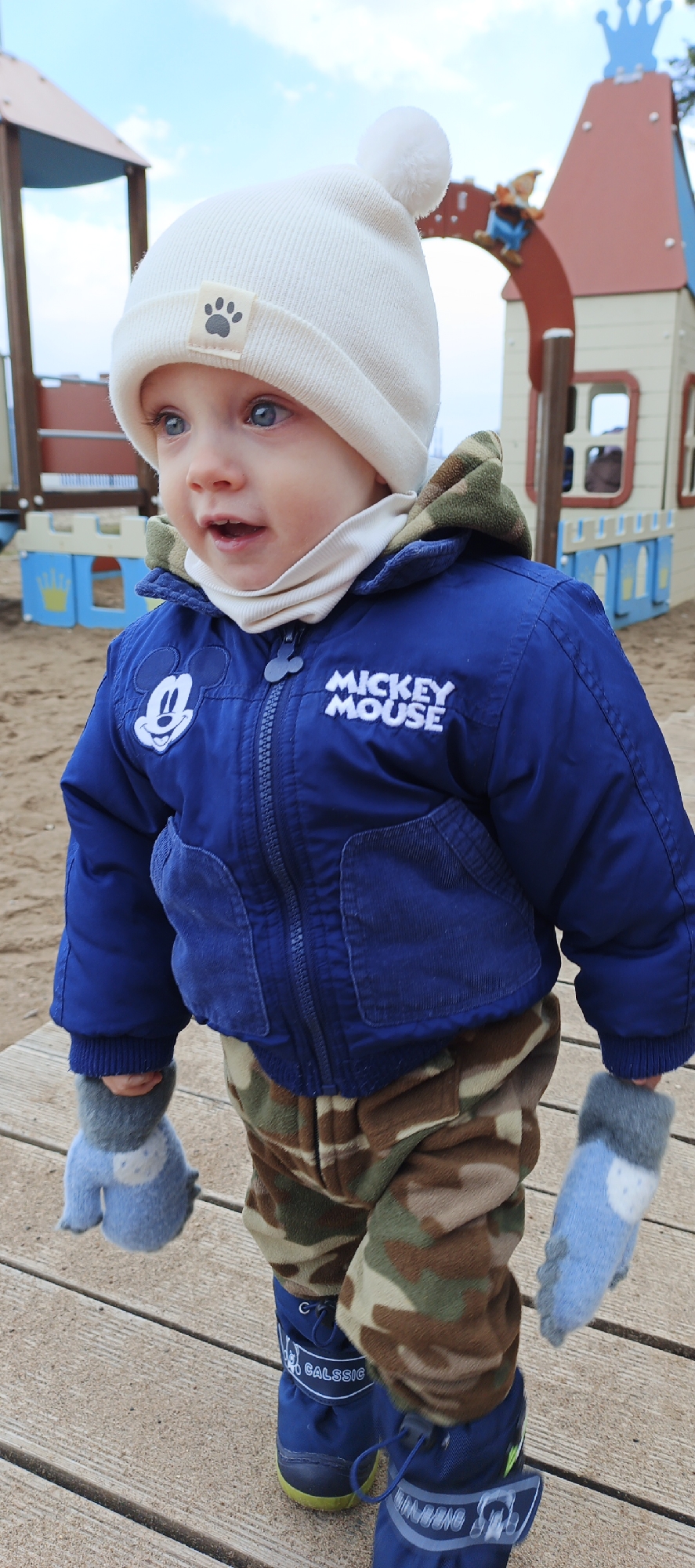A young child stands outdoors on a wooden surface, likely a deck or platform, in Chaikovsky, Russia. The child is wearing a white knit hat with a pompom on top and a paw print emblem, a blue jacket adorned with Mickey Mouse imagery, camouflage-patterned pants, and blue snow boots. They also have light blue and gray mittens on. The backdrop reveals a playground structure with a castle-like appearance. The sky is visible above the structure. The overall setting suggests a public park or recreational area.