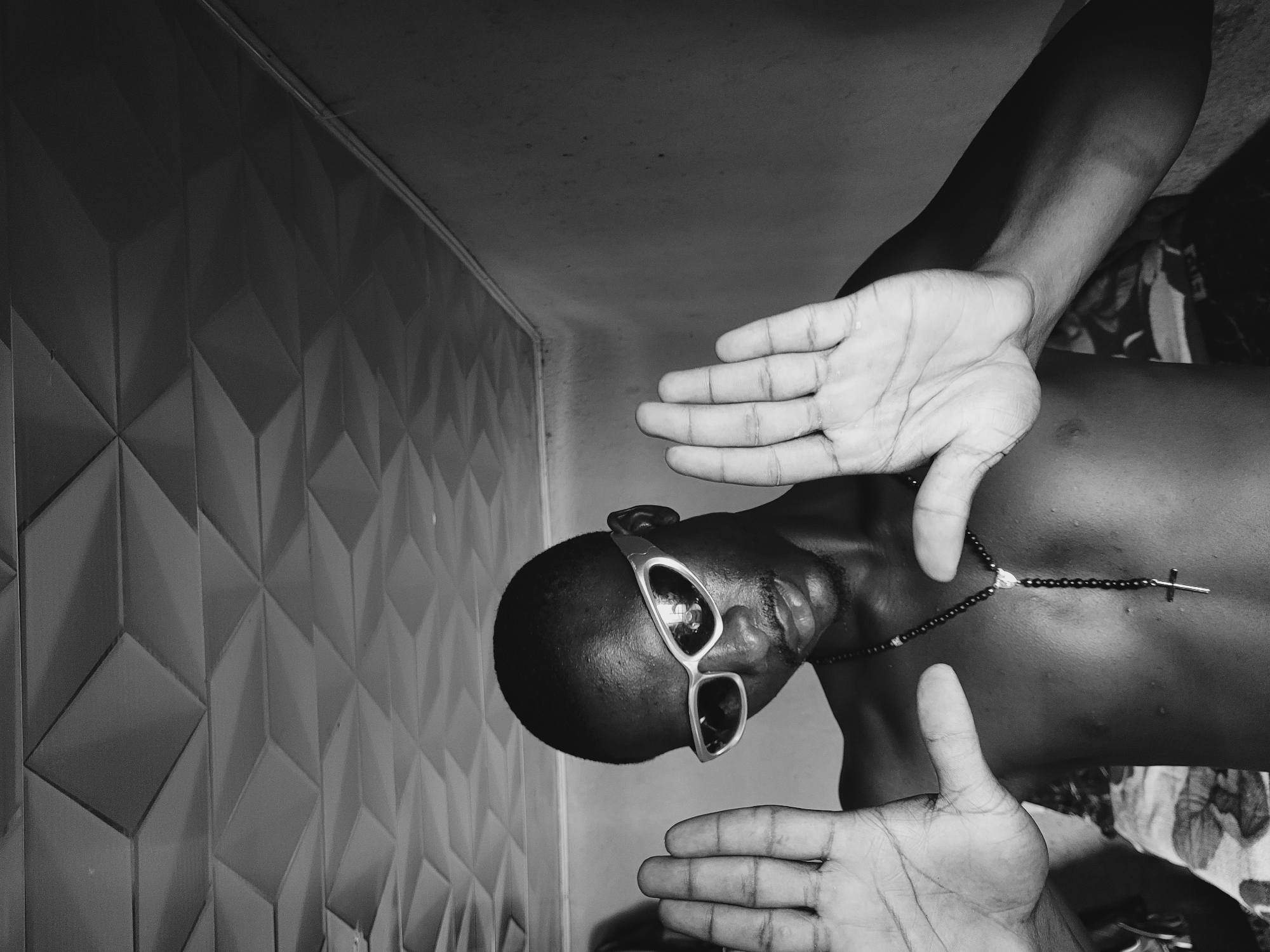 The monochrome image depicts a person with dark skin, wearing sunglasses and a beaded necklace with a cross pendant, framed by their own hands. The person's head is angled downwards. The background includes a textured wall with geometric patterns and a ceiling. The specific location and city of Bukuru, Nigeria, are not discernible from the image alone.