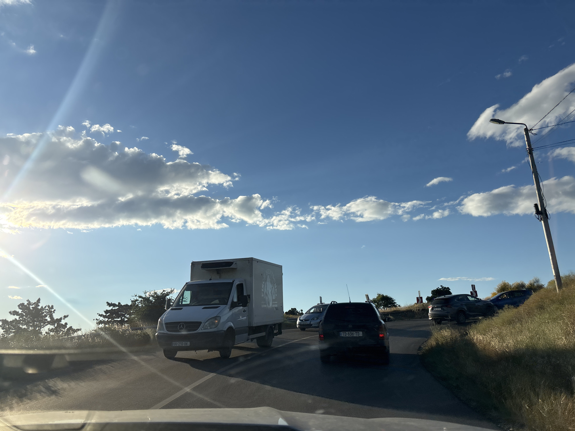 The image captures a street view in Tbilisi, Georgia, under a bright, partly cloudy sky, likely late afternoon due to the angled sunlight casting long shadows and creating lens flares. The scene depicts a road with several vehicles, suggesting traffic.
In the foreground, a white refrigerated truck with "PULKOVO" visible on its side is moving along the road. To its right, a dark-colored SUV with license plate "TD 836-TD" is also in motion. Further ahead and to the right, a dark grey SUV is partially off the road, on a grassy embankment, with a blue car behind it, possibly involved in an incident or parked in an unusual manner. Several red and white directional signs are visible along the roadside, indicating a curve in the road.
The sky is a vibrant blue, punctuated by fluffy white clouds, with a noticeable sunbeam cutting across the upper left portion of the frame. A lamppost with electrical wires stands on the right side of the road, characteristic of urban or suburban street lighting. Greenery, including trees and tall grass, lines the sides of the road. The perspective suggests the photo is taken from inside another vehicle, looking forward.
No people are clearly visible within the frame. The overall mood is that of an ordinary day on the road, with the potential for a minor traffic event implied by the SUV on the embankment.