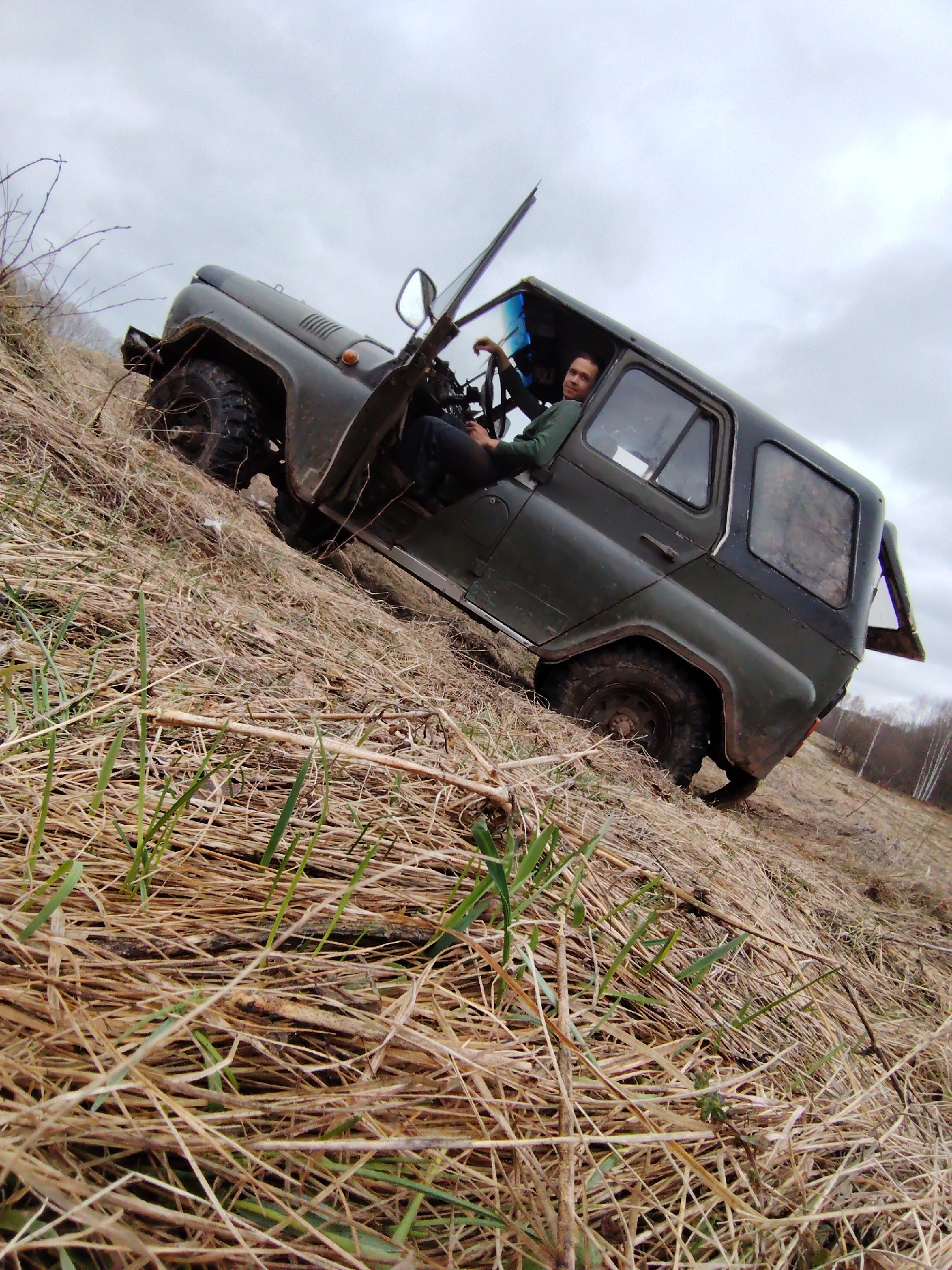 An off-road vehicle, dark green and somewhat muddy, is dramatically tilted on a steep, grassy slope under an overcast sky. A man, wearing a green shirt, is seated in the driver's seat with the door open, looking towards the camera with a relaxed, slightly smiling expression.
The scene is an unpaved, rural landscape, possibly near Markovskii, Russia, featuring a sharp incline covered in a mix of dry, straw-like grass and patches of new green growth, suggesting early spring or late autumn. The vehicle's rugged tires are visible, dug into the terrain. In the background, beyond the slope, a line of bare trees can be seen under the uniform grey sky. The photograph is taken from a low angle, enhancing the sense of the vehicle's precarious position and the steepness of the terrain. There is no visible text in the image.