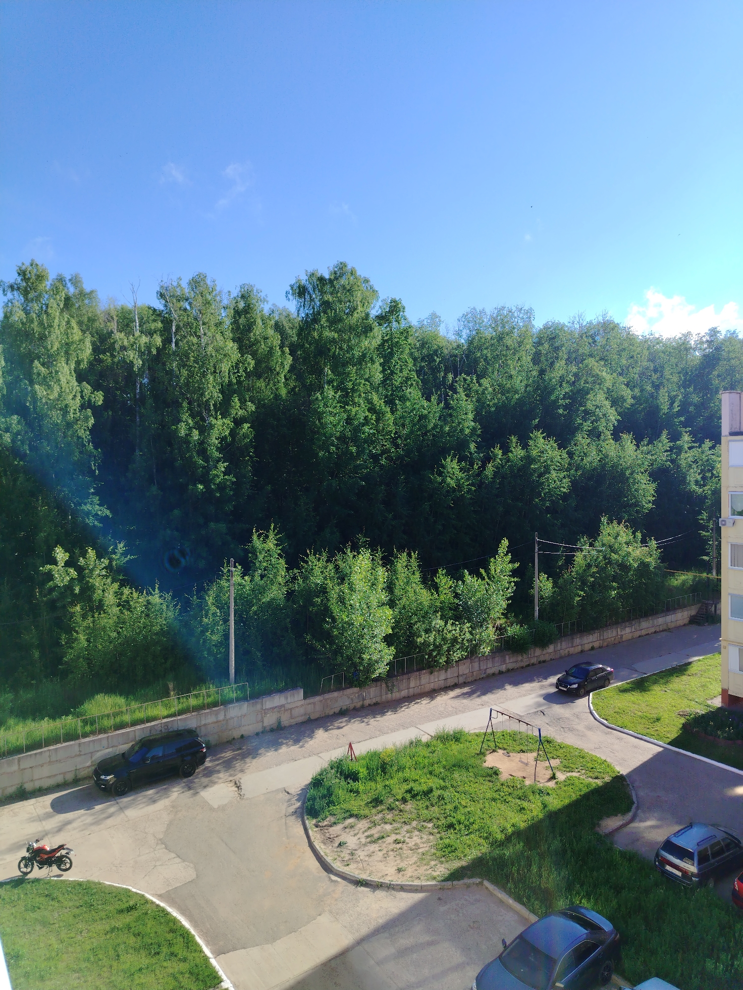 The image depicts a scene in Noviy, Russia, under a clear blue sky with scattered clouds. In the foreground, a residential building with a light yellow facade is visible on the right side. A paved road curves through the scene, bordered by concrete barriers and grass. Several vehicles are present: a black SUV parked on the side of the road, a motorcycle parked on a grassy area, a dark sedan driving on the road, and two other cars parked in a lot near the building. A children's swing set with two swings stands on a patch of grass between the road and the forest. A dense forest with lush green trees occupies the background, forming a natural boundary. A few utility poles with wires are also visible.