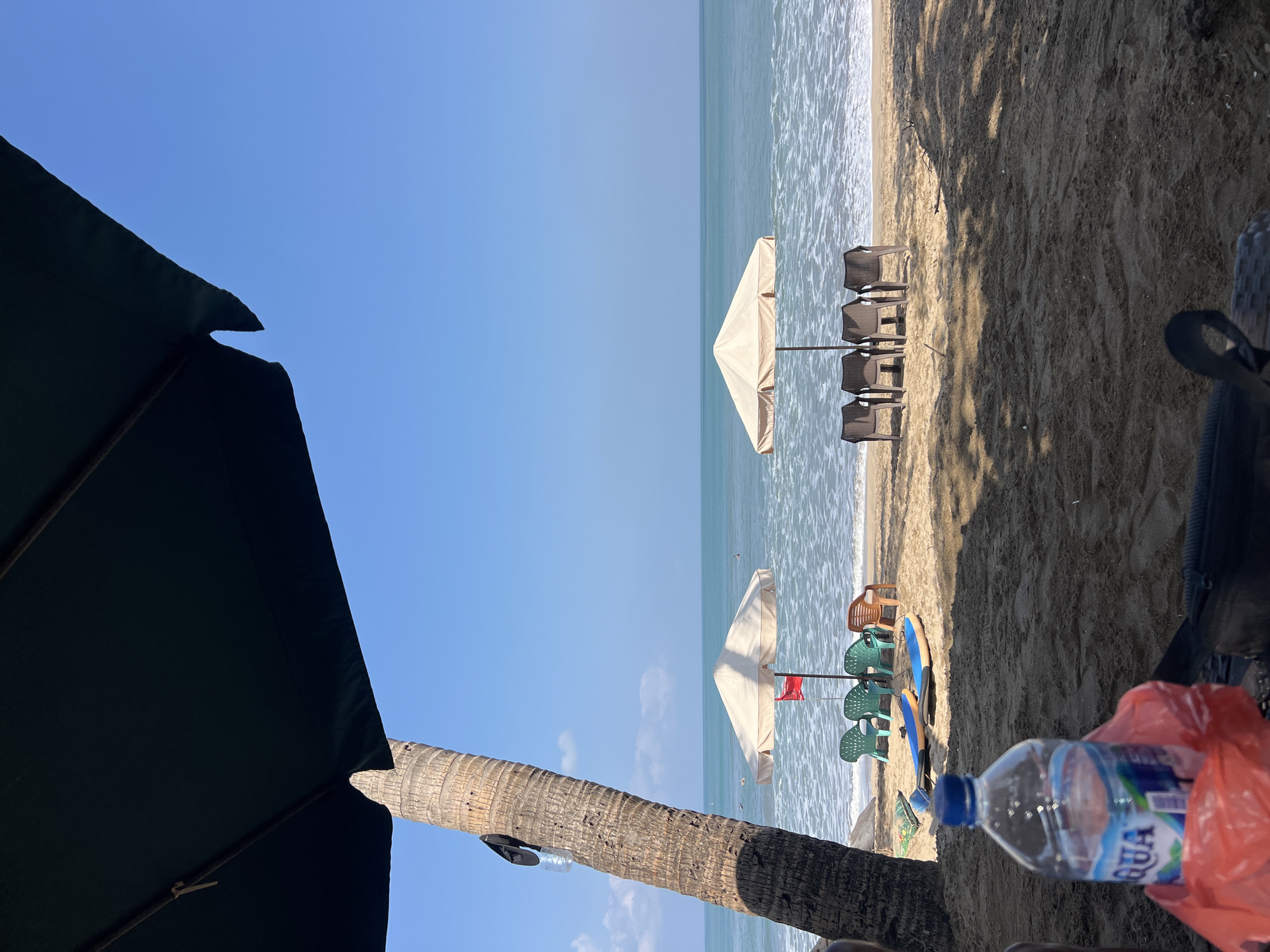 A beach scene in Kuta, Indonesia, is captured under the shade of a large dark umbrella and a palm tree on the left. The sand is textured with visible footprints and the shadows of overhead objects. In the foreground, a clear plastic bottle of Aqua bottled water is placed on top of a red plastic bag. Beside it, a black bag rests on the sand.
Further back on the beach, two surfboards with blue tops and white undersides are lying on the sand. Two beach umbrellas stand open, providing shade. The left umbrella is lighter in color and positioned behind several green plastic chairs. A small red flag is visible behind this umbrella. To the right, a second, similar-colored umbrella is set up behind a row of four brown plastic chairs.
The ocean stretches out in the background, with gentle waves breaking on the shore and creating white foam. The water is a vibrant blue under a clear, bright sky with a few wispy clouds. Two figures can be seen in the water, seemingly swimming or wading. The overall atmosphere is that of a tranquil and sunny day at a popular tourist destination.