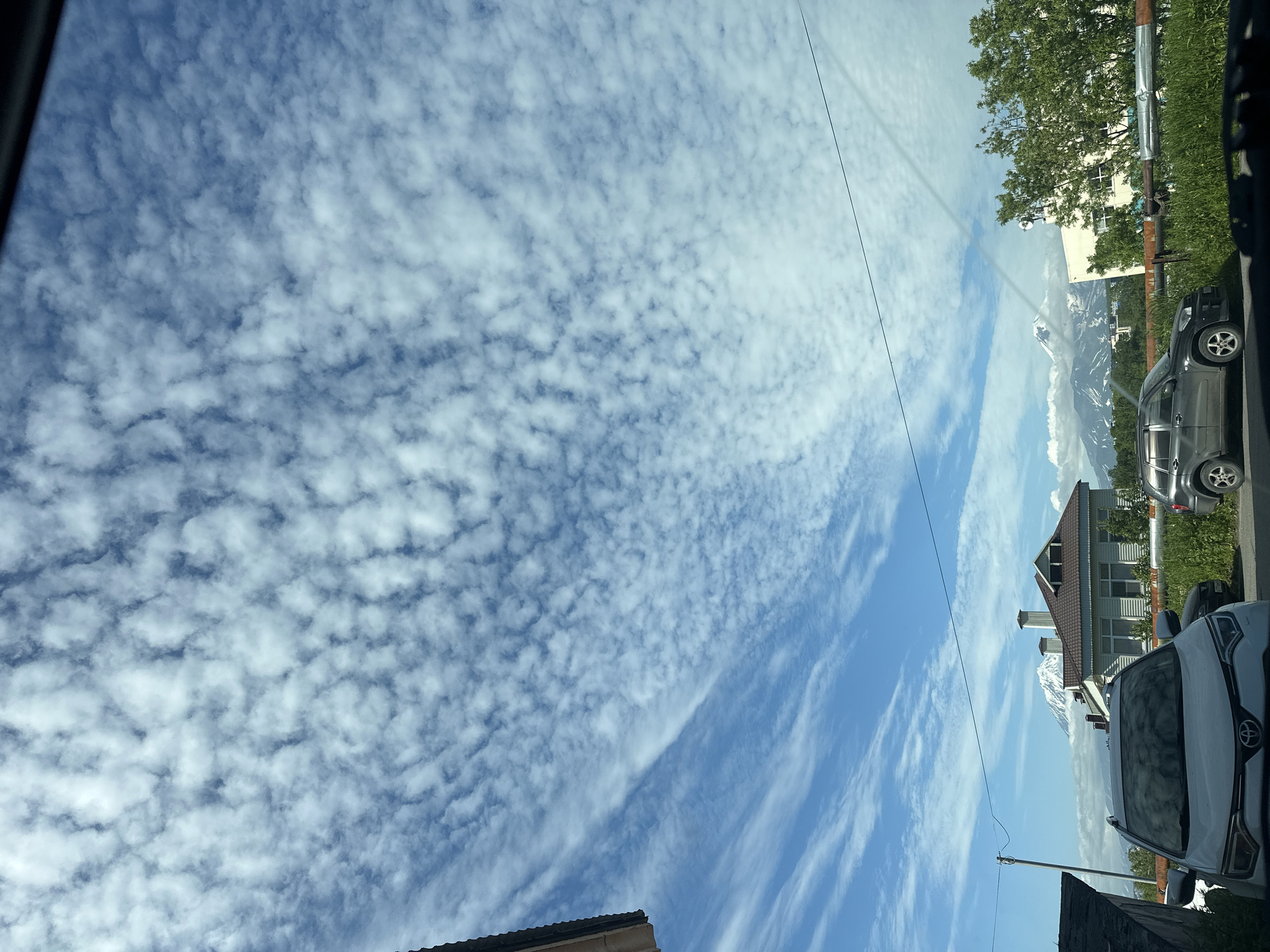 A view from a car window showcases a scene in Petropavlovsk-Kamchatsky, Russia. In the foreground, the hood and windshield of a white Toyota RAV4 are visible, reflecting the sky. To the right, a dark SUV is parked. Beyond the vehicles, a residential house with a brown roof and several chimneys stands, partially obscured by pipes and lush green grass. In the distance, a snow-capped mountain is partially covered by clouds, suggesting a backdrop of volcanic landscapes common to the Kamchatka region. The sky is predominantly blue with a pattern of scattered altocumulus clouds. The overall impression is a snapshot of a residential area with natural volcanic features in the background.