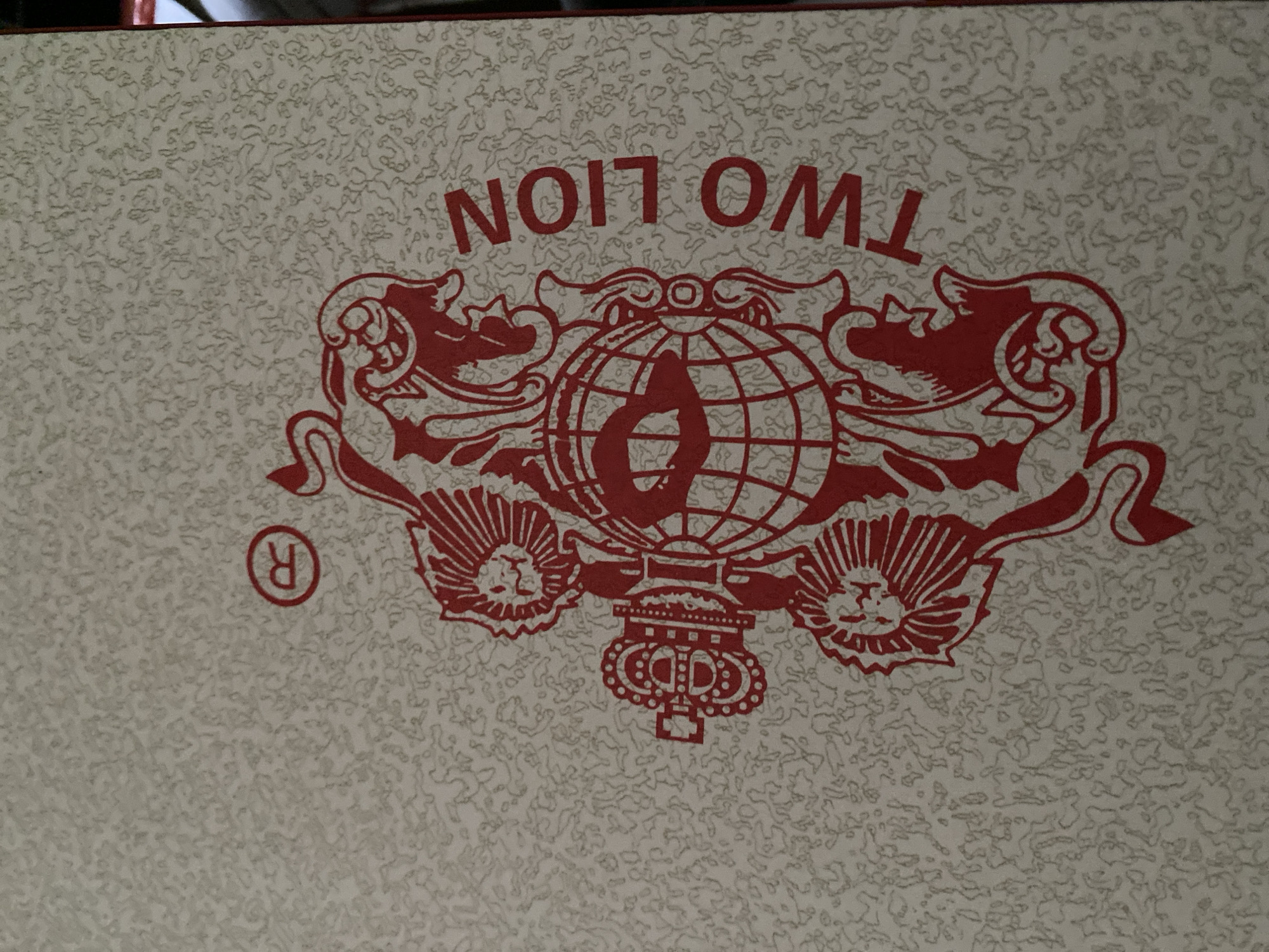This is a close-up, eye-level, vertical shot of a textured, cream-colored surface with a red graphic design. The design features two stylized lions facing each other, with a crown and a globe in between them. The word "TWO LION" is printed vertically along the right side of the graphic in red block letters. A registered trademark symbol (R in a circle) is present in red in the upper left portion of the image. The background has a subtle, organic, mottled pattern in a lighter shade. There is a thin, dark red border along the right edge of the image. This image depicts a graphic design, likely from a product label or packaging, and does not contain any elements related to the city of Jauro Yinu, Nigeria.