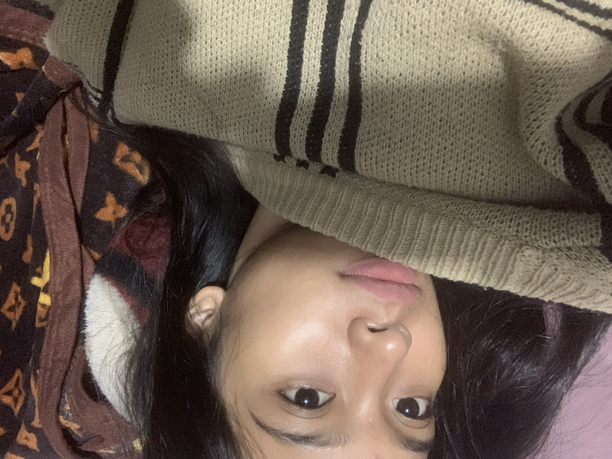 A close-up, inverted selfie of a person with dark hair, light skin, and brown eyes. The person is wearing a beige knit sweater with dark vertical stripes on the right side of the frame. The left side shows part of a brown blanket or jacket with a repeating pattern of stylized flowers or quatrefoils. The background appears to be a solid light purple or pink. The location is not determinable from the visual data.