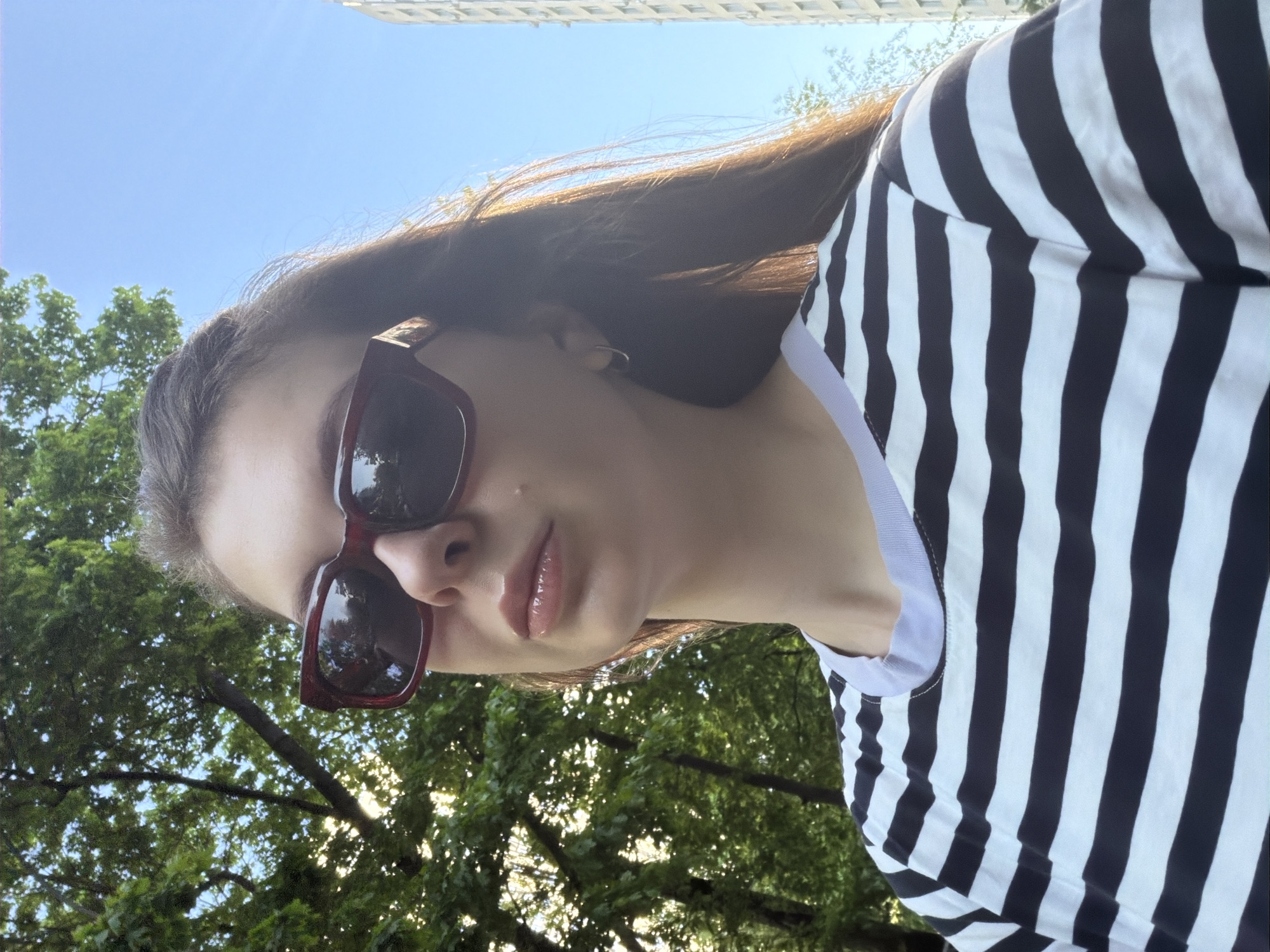 A young adult female is angled downwards towards the viewer, wearing maroon-framed sunglasses and a black and white striped top with a white neckline. She appears to be outside, as evidenced by the trees with green foliage visible in the background, along with patches of blue sky. A partial view of a light-colored building is visible in the upper portion of the frame. This image was taken in Khimki, Russia.