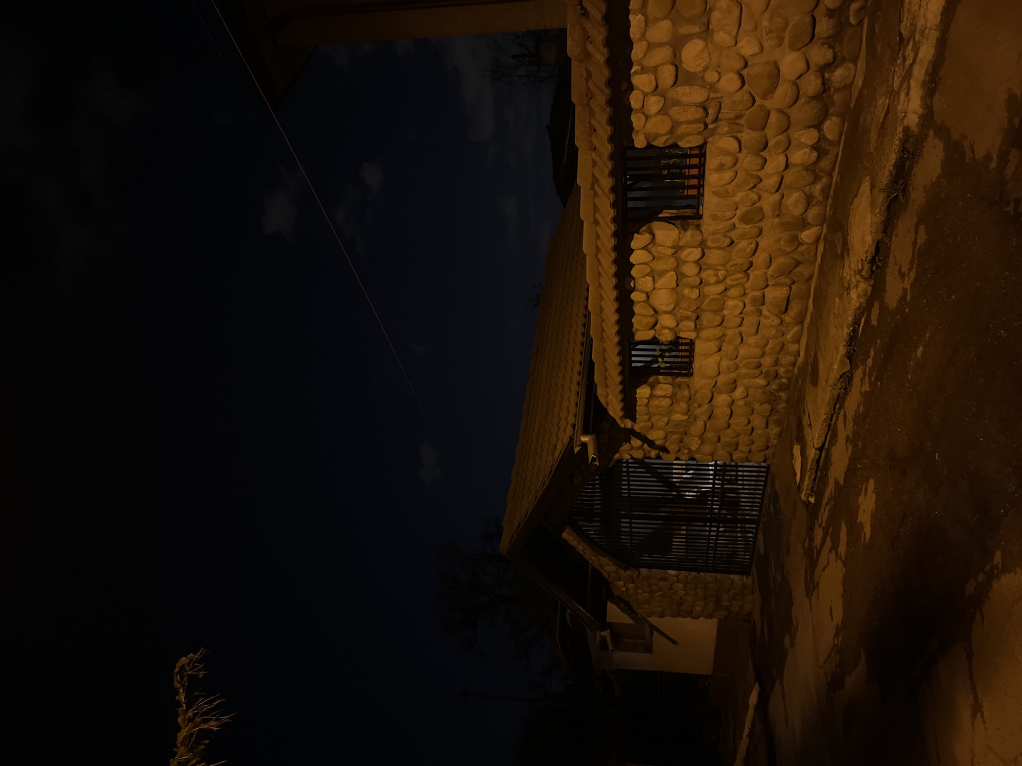 A nighttime shot captures a building with a unique facade. The exterior is constructed from rounded, light-colored stones arranged in a mosaic-like pattern. Windows are set into the stone wall. The sky above the building is dark. A powerline is visible above the roof. The location is likely Sofia, Bulgaria.