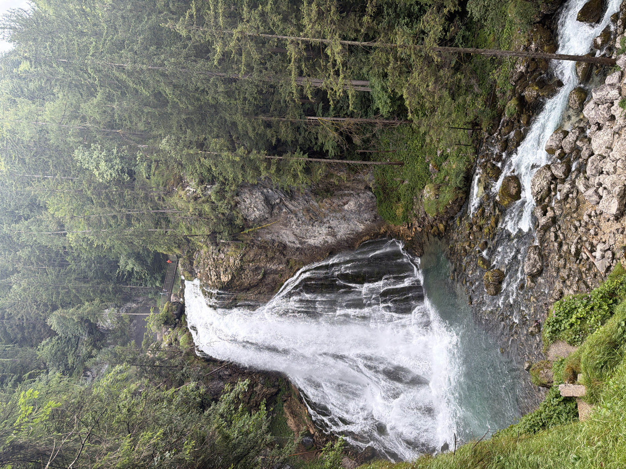 This media file shows a powerful waterfall cascading down a rocky cliff face in a lush, forested mountain environment. Tall, dense evergreen trees dominate the upper portions of the image, creating a natural canopy. The waterfall itself is the central subject, with white water rushing and churning over layered rock formations, eventually pooling into a calm, turquoise body of water at the base. The surrounding terrain is rugged and natural, with rocks, moss, and various green vegetation clinging to the slopes.
In the upper right portion of the scene, a small figure wearing an orange jacket can be seen on a wooden walkway that appears to lead along the cliff edge, offering a vantage point to view the waterfall and the surrounding landscape. This walkway suggests that the location is a tourist destination or a place with developed infrastructure for visitors.
The overall impression is one of natural grandeur and a slightly dramatic, yet serene, outdoor setting. The lighting suggests it might be a cloudy or overcast day, as there are no strong shadows, and the light is diffused, which can contribute to a sense of peacefulness and highlight the vibrant greens of the foliage and the color of the water. The scene is likely captured in the daytime, given the visibility of the details. There is no visible text in the image. The location is identified as Kellau, Austria.