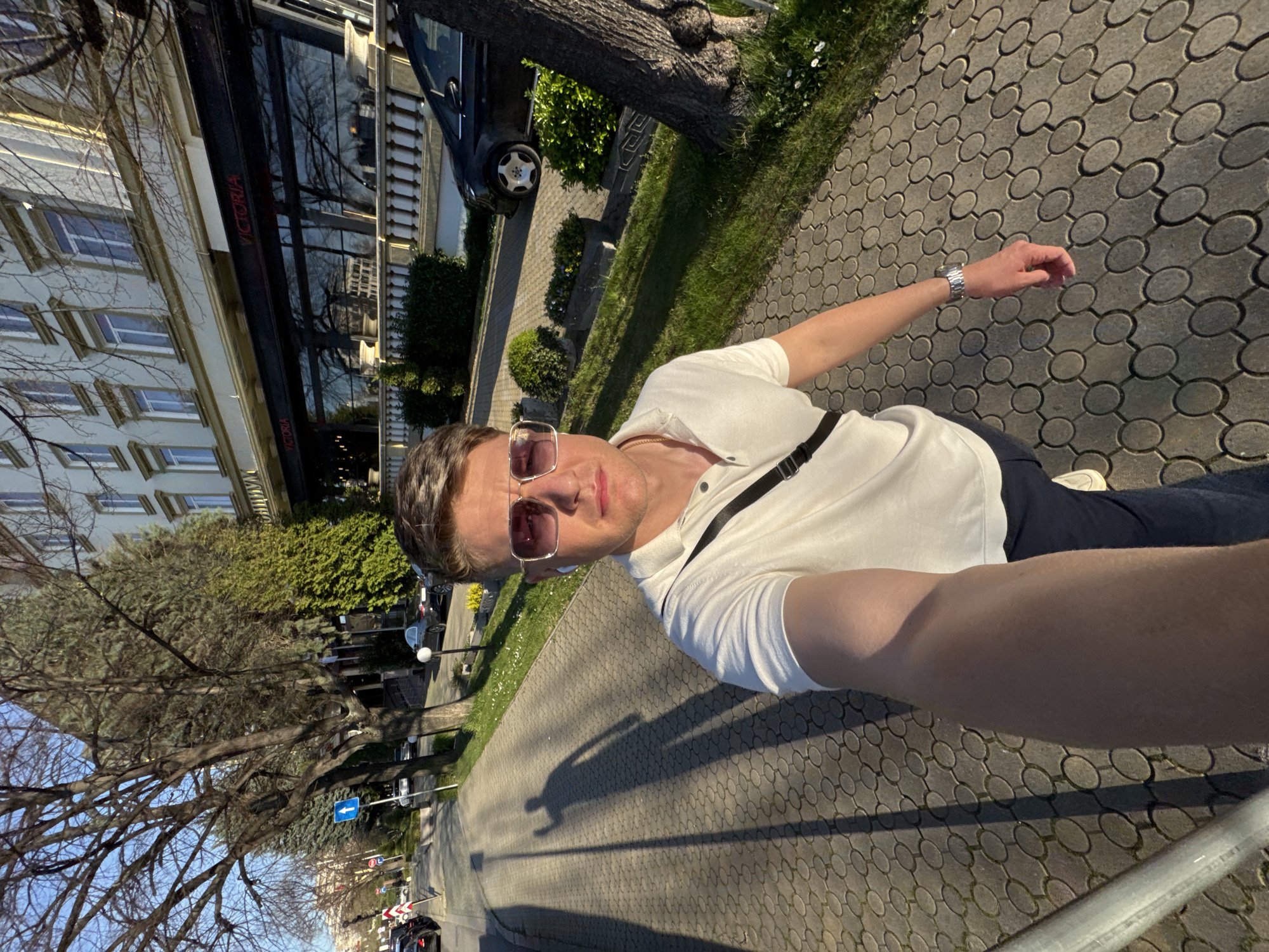 The image depicts a man taking a selfie while standing on a cobblestone sidewalk. He is wearing sunglasses, a light-colored polo shirt, and dark pants. A strap crosses his chest. The sidewalk runs alongside a street with parked cars and trees lining the edge. In the background, there's a building with "VICTORIA" displayed on its facade. The building's architecture suggests an urban setting, and the presence of European street signs in the distance indicate it's a European city. Given the presence of "VICTORIA" on the building, it is likely in Plovdiv, Bulgaria, where the restaurant "Victoria" is located.