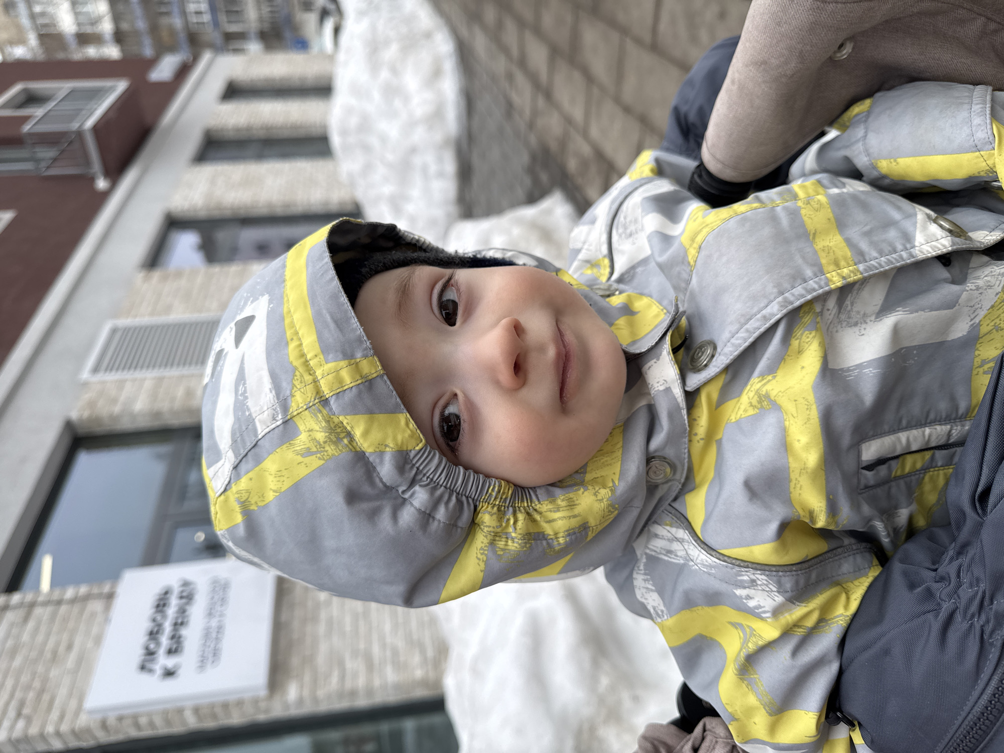 A young child, approximately 1-3 years old, is depicted in a medium close-up, looking directly towards the camera. The child wears a grey and yellow patterned hooded winter suit with a dark beanie visible beneath the hood. The child's dark eyes and a slight smile are visible. The child appears to be seated in a stroller or similar carrier, with dark grey and light brown fabric elements visible in the lower portion of the frame.
The scene is set outdoors in a winter urban environment. In the background, immediately to the child's left, is a building with a light-colored, textured facade. Affixed to this building is a white rectangular sign displaying Cyrillic text: "ЛЮБОВЬ К БРЕДУ" (Lyubov' k Bredu), with smaller, less legible text below it. This sign corresponds to a commercial establishment located at Pushkinskaya Street, 276a, Izhevsk, Russia.
Further into the background, snow accumulates in large piles along a paved walkway or sidewalk. Additional multi-story buildings are visible, including one with a reddish-brown upper section and a small balcony, and another multi-unit residential building in the distance. The ground immediately to the child's right consists of paving stones. The overall environment indicates cold weather conditions.