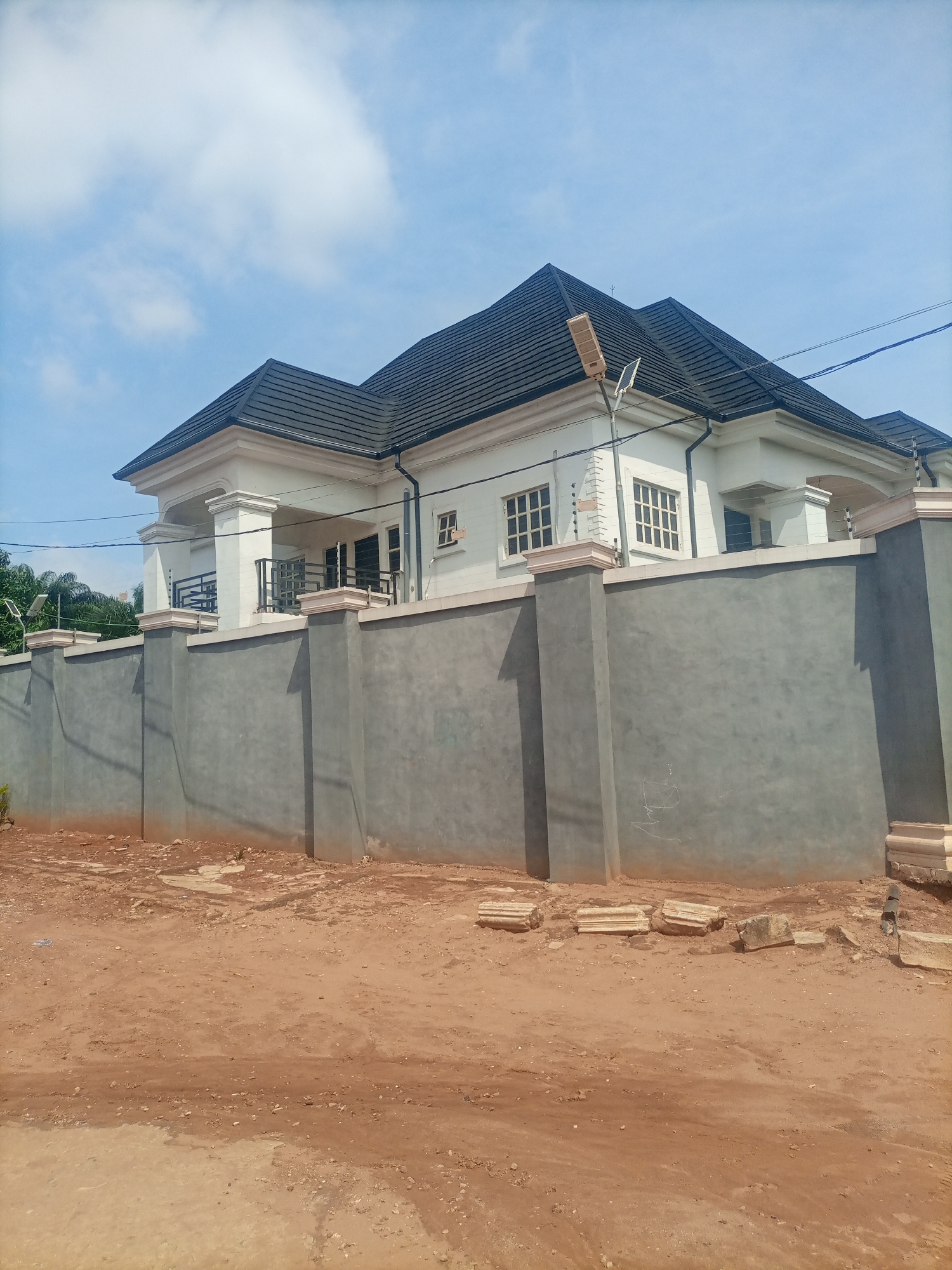 The image depicts a modern two-story house with a dark grey tiled roof, white walls, and multiple windows. A tall grey concrete fence encloses the property. A solar-powered street lamp is visible in front of the house. The scene appears to be under construction or recently completed, as there are piles of building materials, including broken concrete pillars, on the ground in front of the fence. The ground is covered in reddish-brown dirt. The sky is clear blue with a few white clouds, suggesting it is daytime. The overall impression is one of a newly built residential property in a developing area. There is no visible text in the image. The location is Nkpor, Nigeria.