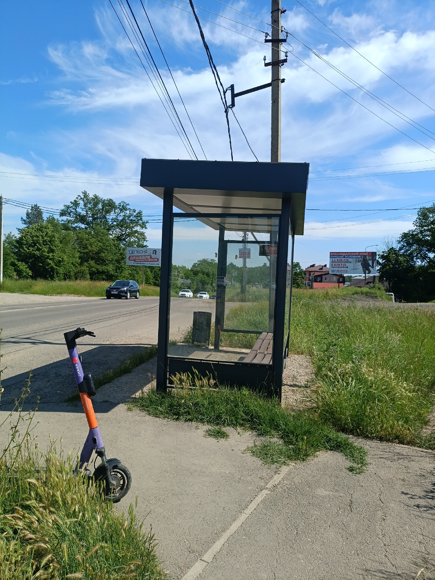 An outdoor scene captures a bus stop shelter with a bench inside, located on the side of a road. A purple and orange electric scooter is parked on the sidewalk next to the shelter. The road is paved and has a white dividing line. Several cars are visible on the road, with one black SUV prominently in the foreground. Lush green grass and trees surround the area. In the background, residential buildings and billboards can be seen. The sky is blue with scattered white clouds. The scene appears to be taken on a bright, sunny day. The billboard in the background advertises "Krasnodar Krai - Best conditions." The location is identified as Goryachiy Klyuch, Russia.