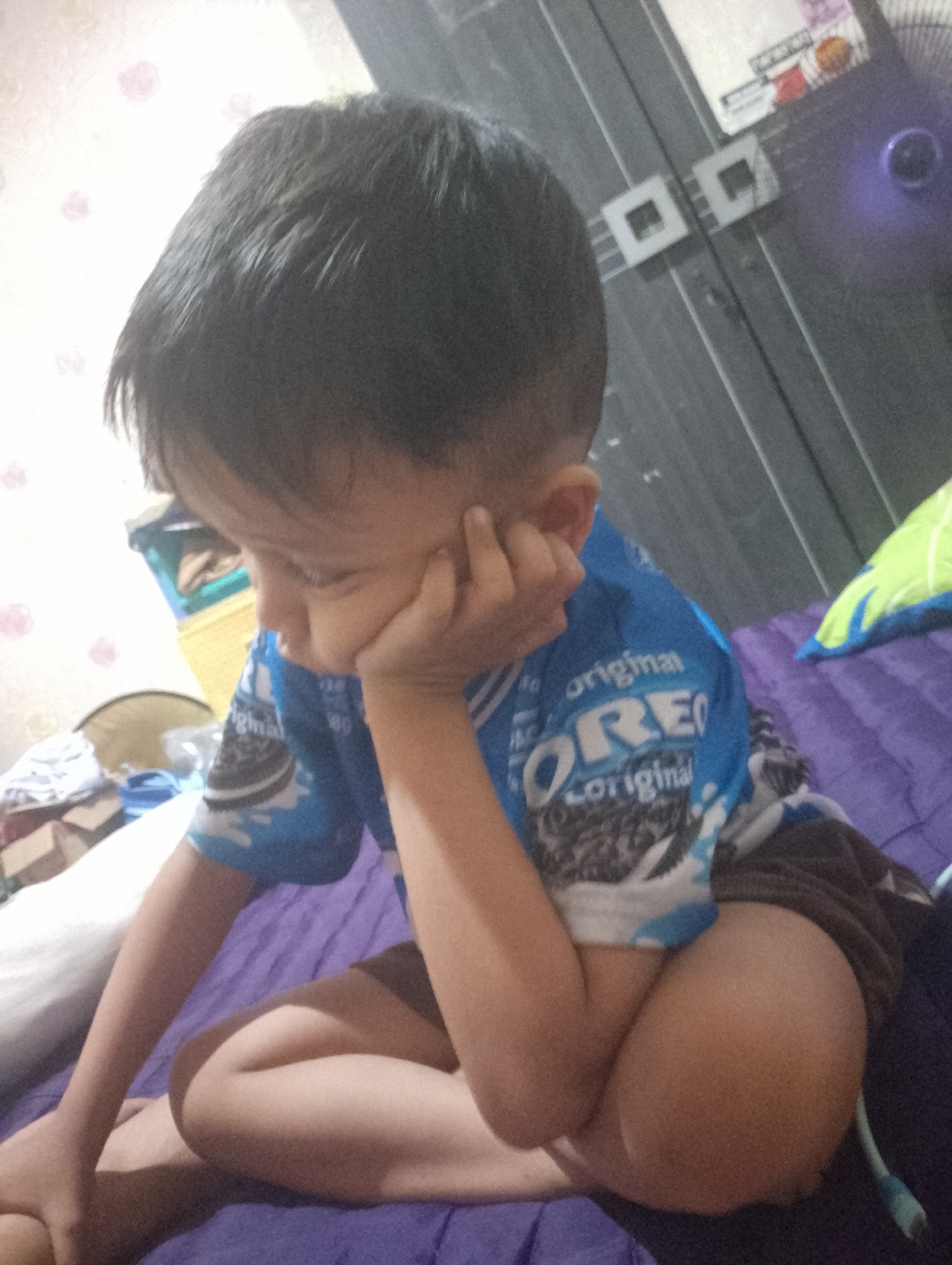 A young boy sits on a purple surface, likely a bed, with his knees bent and his head resting on his hand. He is wearing a blue t-shirt with an Oreo cookie pattern and brown shorts. The t-shirt has the word "original" visible in white text. Behind him, a closet door with two square handles is visible. To the left of the boy, a patterned wallpaper with pink floral designs can be seen, along with some stacked boxes and a round object. A green and blue pillow is on the right side of the image. The lighting suggests it is indoors, possibly in the late afternoon or evening. The boy's posture and expression convey a sense of contemplation or boredom. The setting appears to be a bedroom in a residential home. The text visible on the t-shirt reads "original" and displays the Oreo logo.