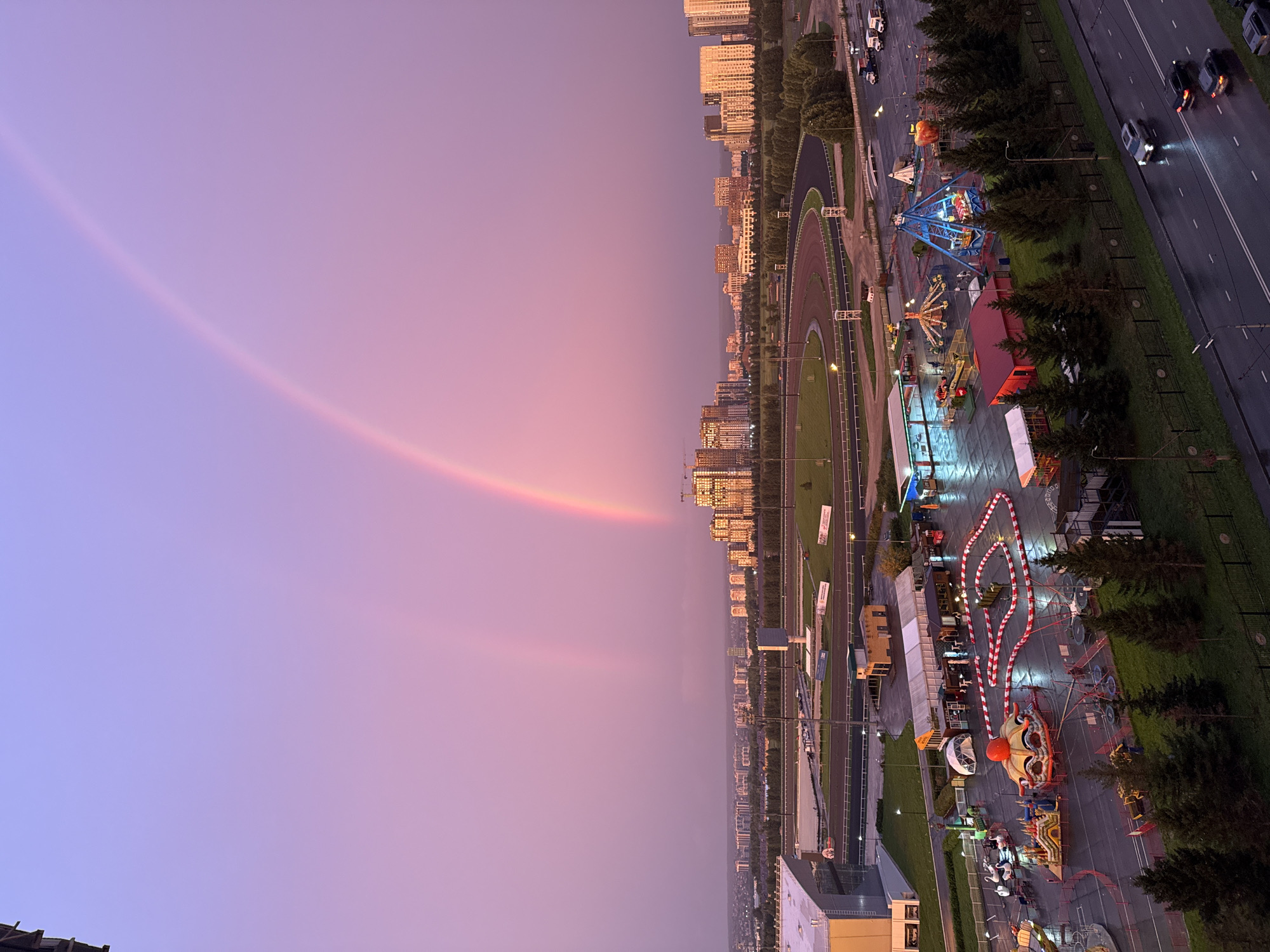 The image captures a panoramic view of a city at dusk, characterized by a vibrant purple and pink sky. A prominent double rainbow arcs across the sky, with its bright colors contrasting against the pastel hues. Below the sky, a cityscape is visible, featuring a race track and several buildings, including tall residential towers. In the foreground, an amusement park is set up with various rides and attractions, including a go-kart track, a carousel, and a swing ride. The ground in the amusement park appears wet, suggesting recent rainfall. Several cars are seen driving on a wet road in the foreground. The overall scene depicts a lively urban environment illuminated by the soft light of dusk and the ethereal glow of a double rainbow.