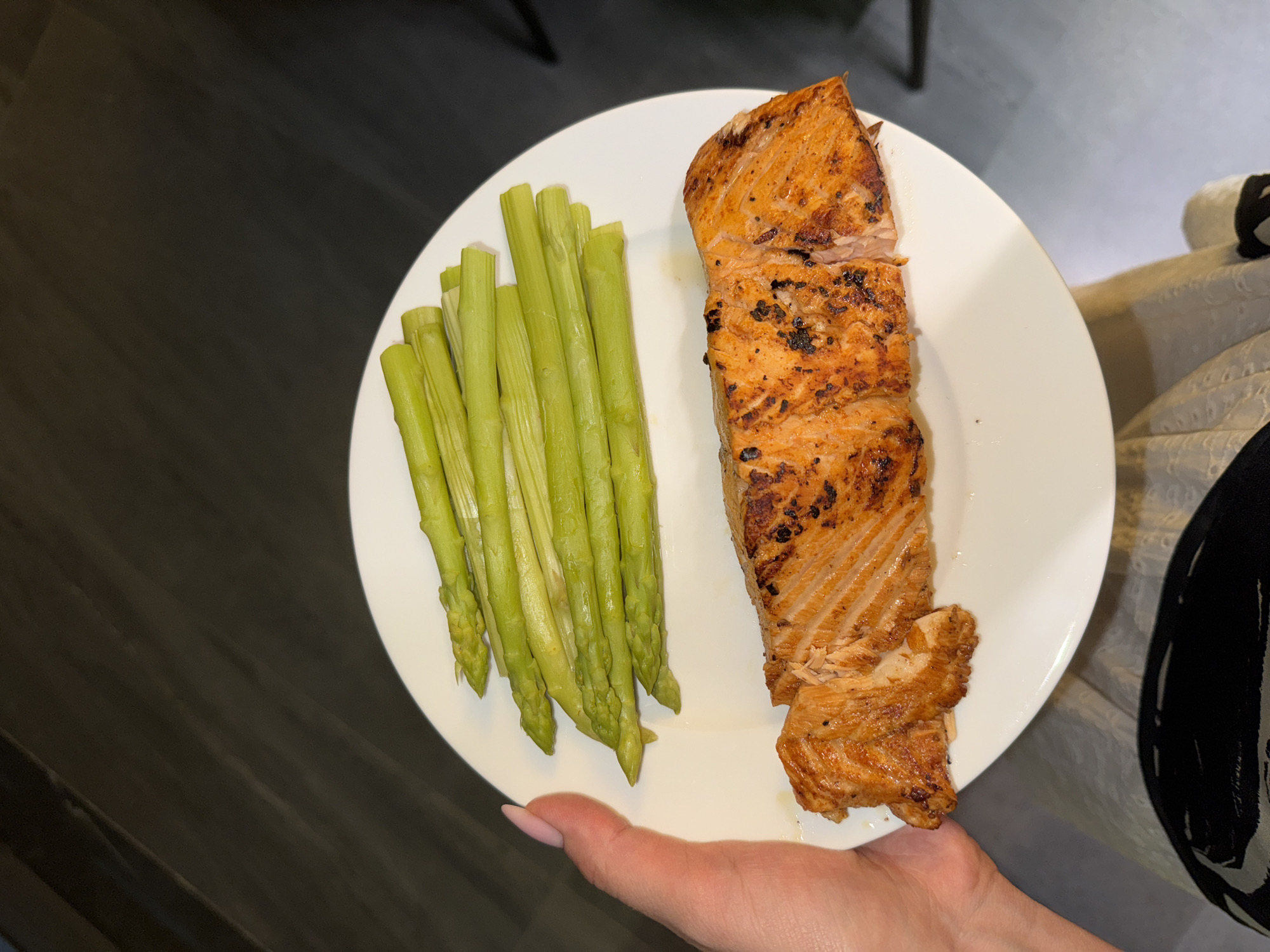 A white plate is held in a person's hand. On the plate, there is a portion of cooked salmon and a bundle of green asparagus spears. The salmon is browned and appears to be grilled. The background is out of focus but shows a neutral-toned surface and fabric, possibly a table and clothing. The location is unspecified.