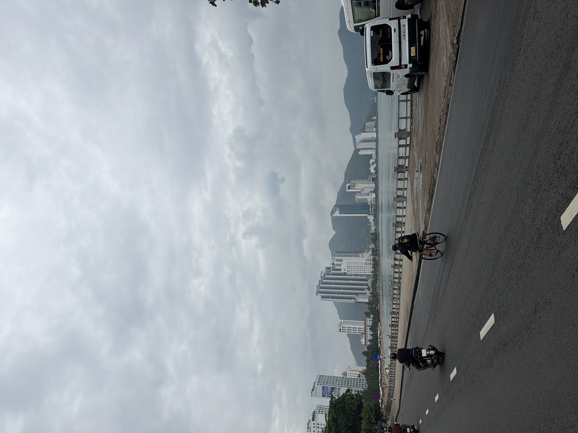 This image depicts a coastal urban scene in Bac Nha Trang Ward, Vietnam, under an overcast sky. The photograph is taken from a slightly elevated perspective, looking along a two-lane asphalt road.
On the road, daily activity is visible, with individuals riding a bicycle and a motorbike in the foreground, traveling away from the camera. Further down the road, additional motorbikes and pedestrians can be discerned. To the immediate left of the road is a concrete barrier, beyond which lies the sea.
The coastline is densely populated with numerous modern high-rise buildings, indicative of a developed urban area. In the background, a range of mountains rises prominently, their peaks partially obscured by the heavy cloud cover that dominates the upper portion of the frame. The sky is a uniform light grey, suggesting a fully cloudy day.
Two white vans are parked on the side of the road near the barrier. On the rear of the closer van, yellow text "TRÂN ĐÀO" is visible, along with a license plate that reads "47H1-054.20". The overall impression is of a bustling coastal city on a cloudy, daytime.