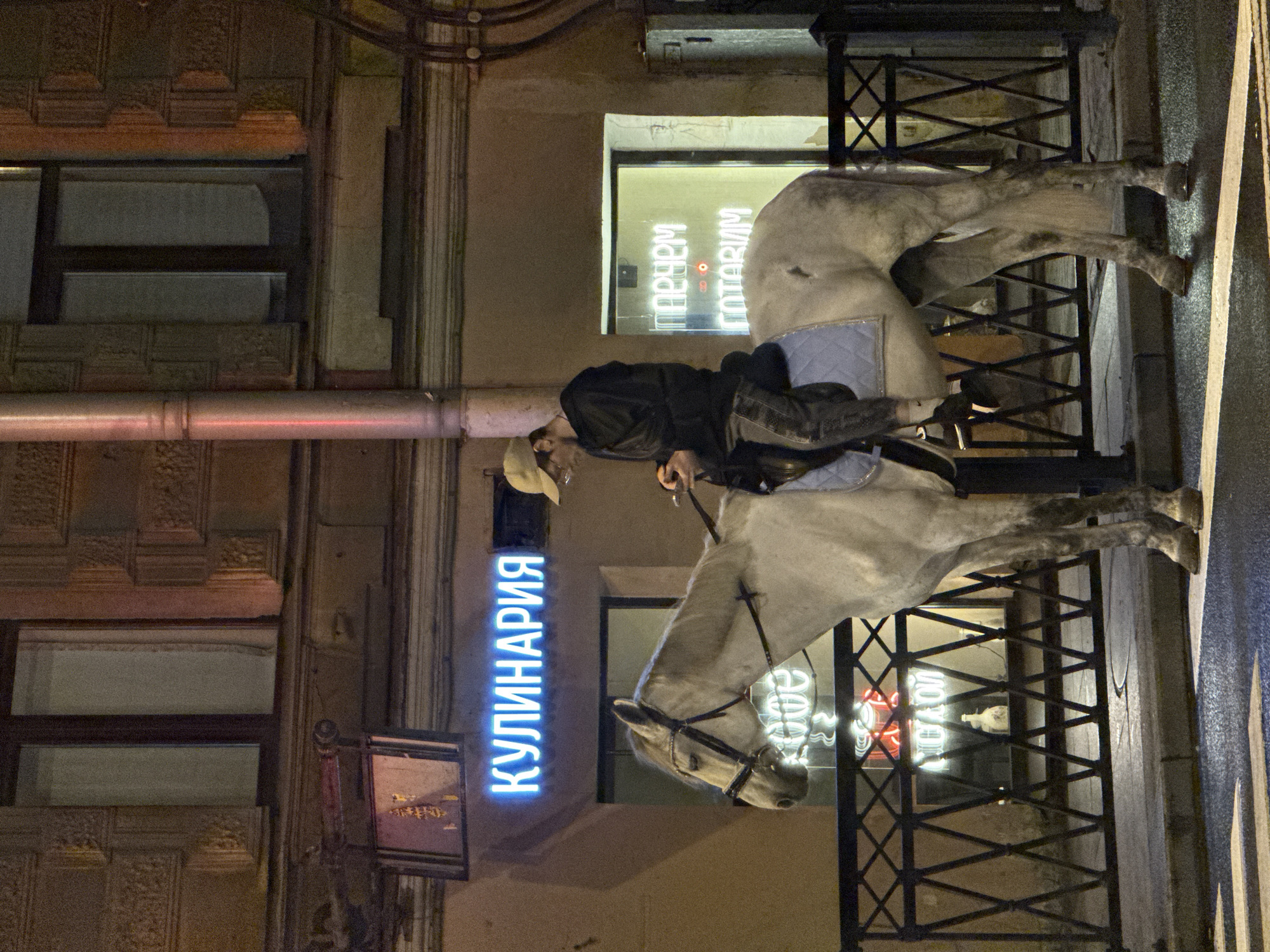 A person wearing a tan baseball cap and a dark hooded jacket sits astride a light-colored horse on a street in Saint Petersburg, Russia. The person is looking down at a smartphone in their hands. The horse is wearing a bridle and a light blue saddle pad. Behind the horse and rider, a neon sign with Cyrillic letters reads "КУЛИНАРИЯ" (KULINARIYA), which translates to "DELICATESSEN." Further back, another neon sign reads "ПАРЧЕМ ГОТОВИМ" (PARCHEM GOTOVIM), which translates to "WE COOK." The street is paved, and there is a metal fence with a diamond pattern separating the sidewalk from a building. The building has decorative architectural details and windows. The scene is illuminated by streetlights and the neon signs, indicating it is nighttime. The overall impression is a surreal urban scene, with the horse and rider juxtaposed against the backdrop of a functioning, illuminated business establishment.
