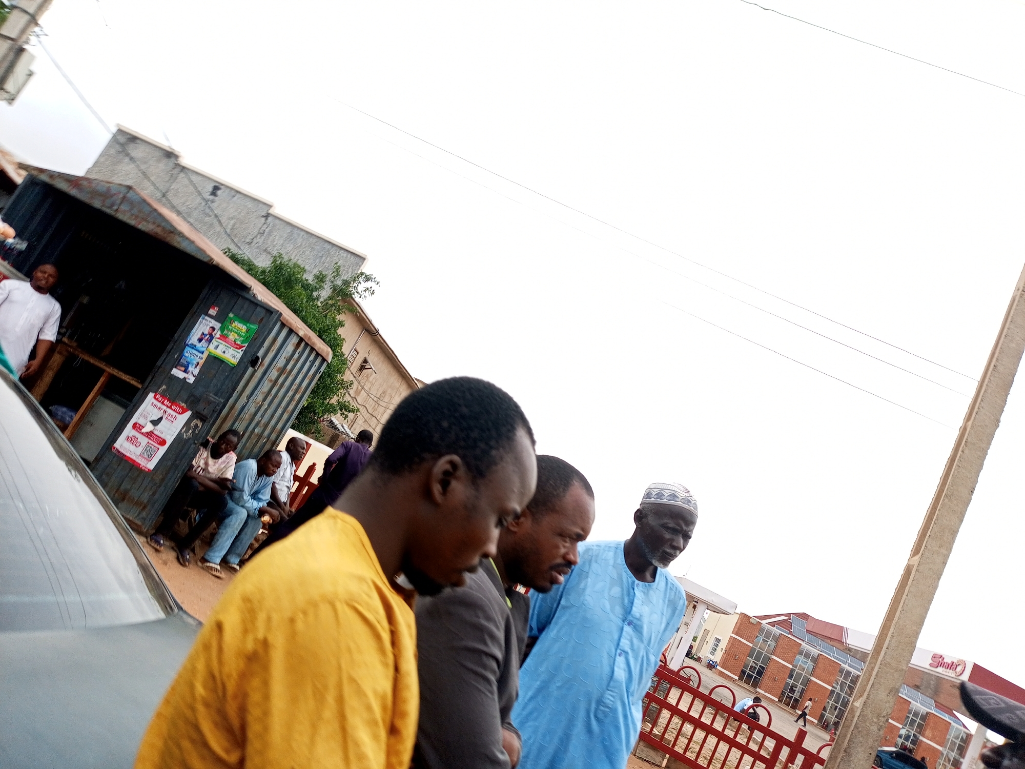 In Tumfure, Nigeria, on a cloudy day, a group of men stand in front of a corrugated metal structure that appears to be a shop or stall. The structure has posters on its exterior. In the foreground, three men are visible from the chest up. The man on the left wears a yellow shirt, the middle man wears a dark grey suit jacket over a lighter shirt, and the man on the right wears a light blue kaftan and a white and grey patterned cap. The man on the right is looking down and to his left, while the other two men are looking in the same direction. In the background, a silver car is partially visible. Further back, several other men are seated and standing near the corrugated structure. The environment appears to be an outdoor, urban setting with buildings and utility poles visible. The overall impression is one of a street scene with people gathered.