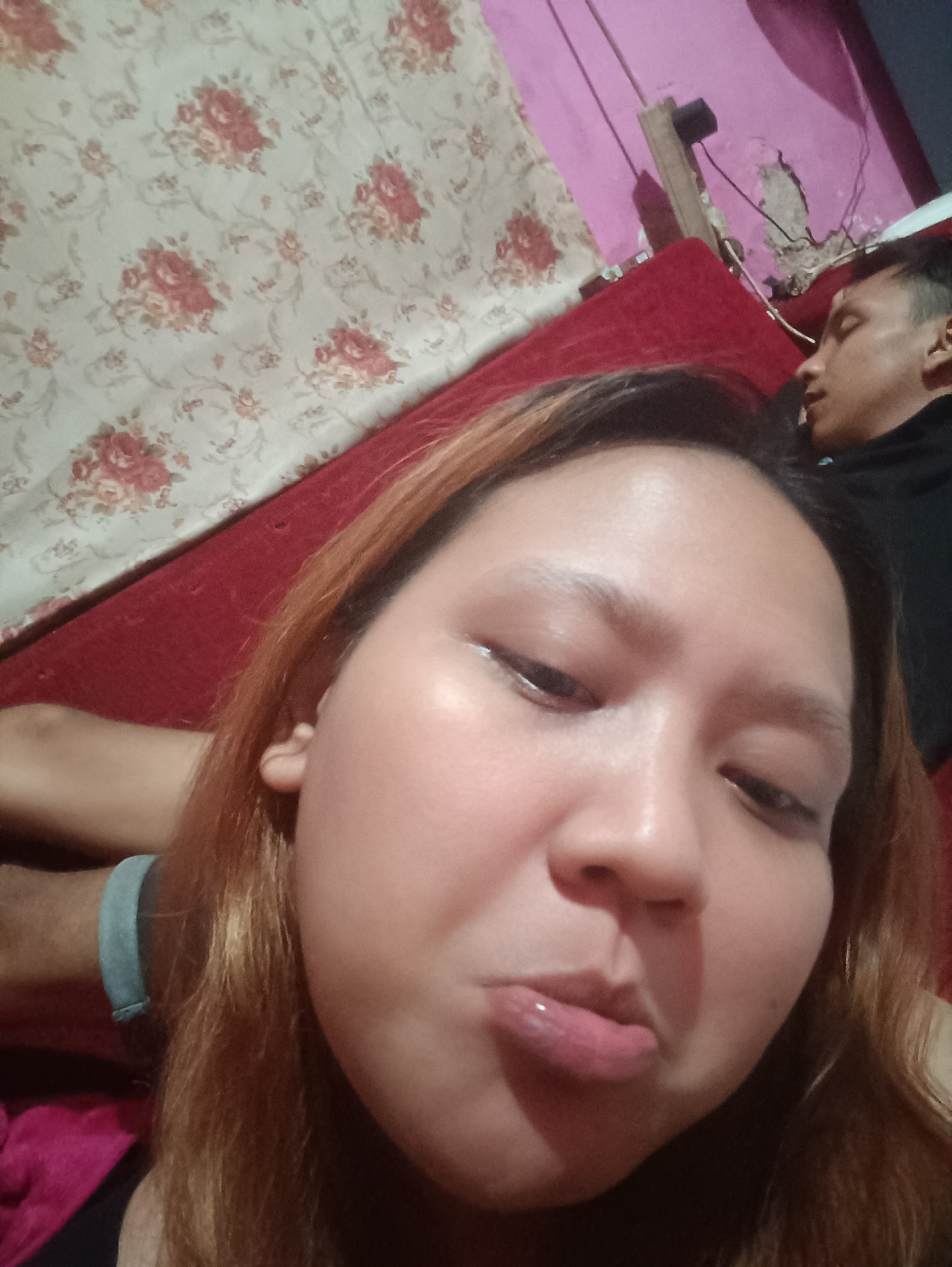 This is a selfie taken by a young woman in Depok, Indonesia, likely in a residential setting, possibly a bedroom. The woman is in the foreground, with her face close to the camera, making a puckered expression. She has long, reddish-brown hair and appears to be wearing lip gloss. Behind her, a man is visible, lying down and seemingly asleep. He is wearing a black shirt. The background includes a red couch, a floral-patterned curtain or fabric, and a pink wall with some visible damage and electrical components. The lighting suggests it is nighttime or indoors with artificial lighting. The overall impression is an intimate, casual moment captured at home.