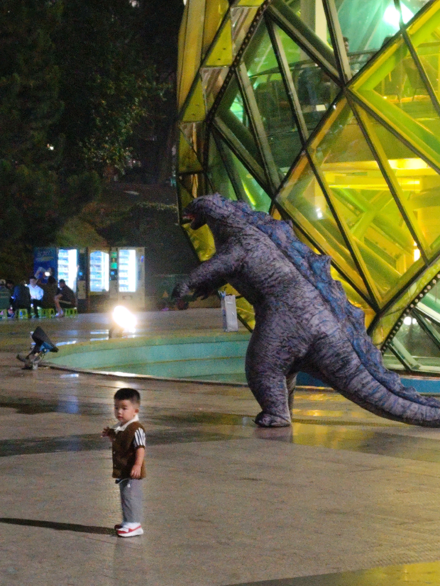The image captures a scene at night in Da Lat, Vietnam, specifically near the iconic glass dome-shaped structure of Cafe Tung. A young boy stands in the foreground, facing the camera. He wears a brown vest over a striped shirt, gray pants, and red and white sneakers. Behind him and to his right is a life-sized statue of Godzilla, positioned as if emerging from or interacting with the glass dome. In the background, vending machines are illuminated, and people are sitting at tables nearby. The ground appears wet, reflecting the surrounding lights. The environment suggests a public space or park area, possibly a popular gathering spot.