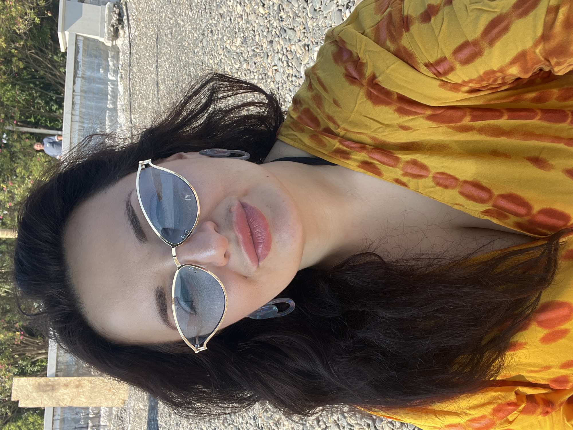 A woman is taking a selfie on a pebble beach with trees and a wall in the background. She has dark, wavy hair and is wearing large, gold-rimmed sunglasses. Her lips are painted with a light pink lipstick, and she is wearing large, translucent earrings. Her top is a yellow and orange tie-dye pattern, and a black bikini top is visible underneath. In the background, a man in a blue shirt is standing on the wall. The scene appears to be a public outdoor space, possibly a park or a waterfront area in Sokhumi, Abkhazia.