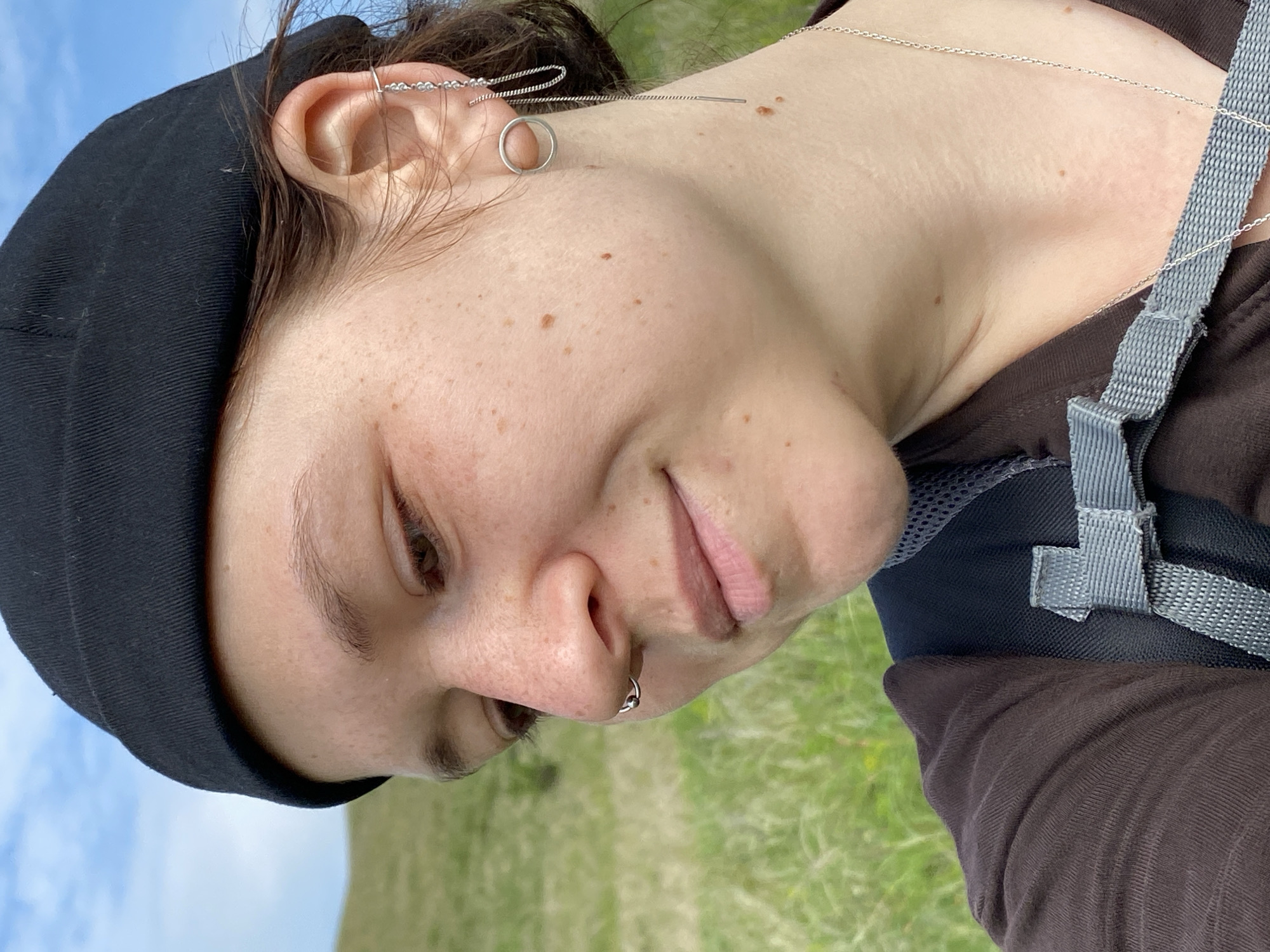 In a close-up, profile shot, a person wearing a black beanie and a brown t-shirt is seen outdoors with a blurred grassy hillside and blue sky in the background. The person has light skin with freckles, a septum piercing, and multiple earrings in their left ear, including a hoop and a chain earring. They are wearing a backpack with grey straps. The image's lighting suggests it was taken during the daytime. The location is Bogoslovka, Russia.