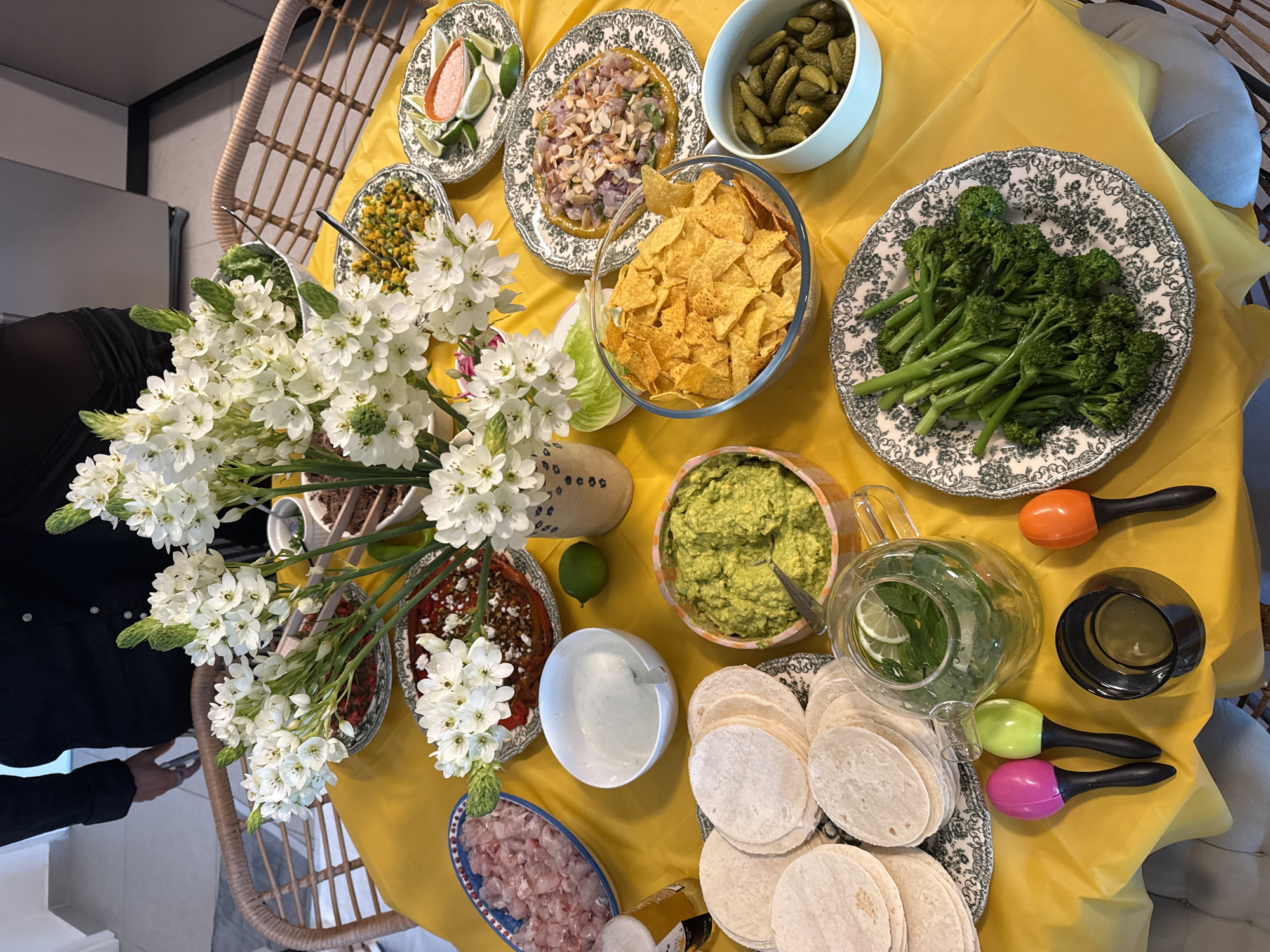 An overhead, close-up shot shows a table laid out with food and decorations, likely in Tel-Aviv, Israel, given the context. A bright yellow tablecloth covers the surface of the table. Dominating the center of the frame is a large floral arrangement of white flowers with green buds, set in a patterned vase. Surrounding the flowers are various dishes of food, including a bowl of guacamole, a bowl of tortilla chips, a plate of corn salsa, a plate of chopped chicken, a plate of what appears to be ceviche with sliced almonds, a plate of pickles, a plate of broccoli rabe, a bowl of dipping sauce, and a stack of small tortillas. A glass teapot with mint and lime slices is also visible, along with a glass of beer, a black candle holder, and three colorful maracas (pink, green, and orange). In the background, a person wearing a black shirt is partially visible, standing behind a rattan chair. The overall impression is of a festive gathering or meal preparation.