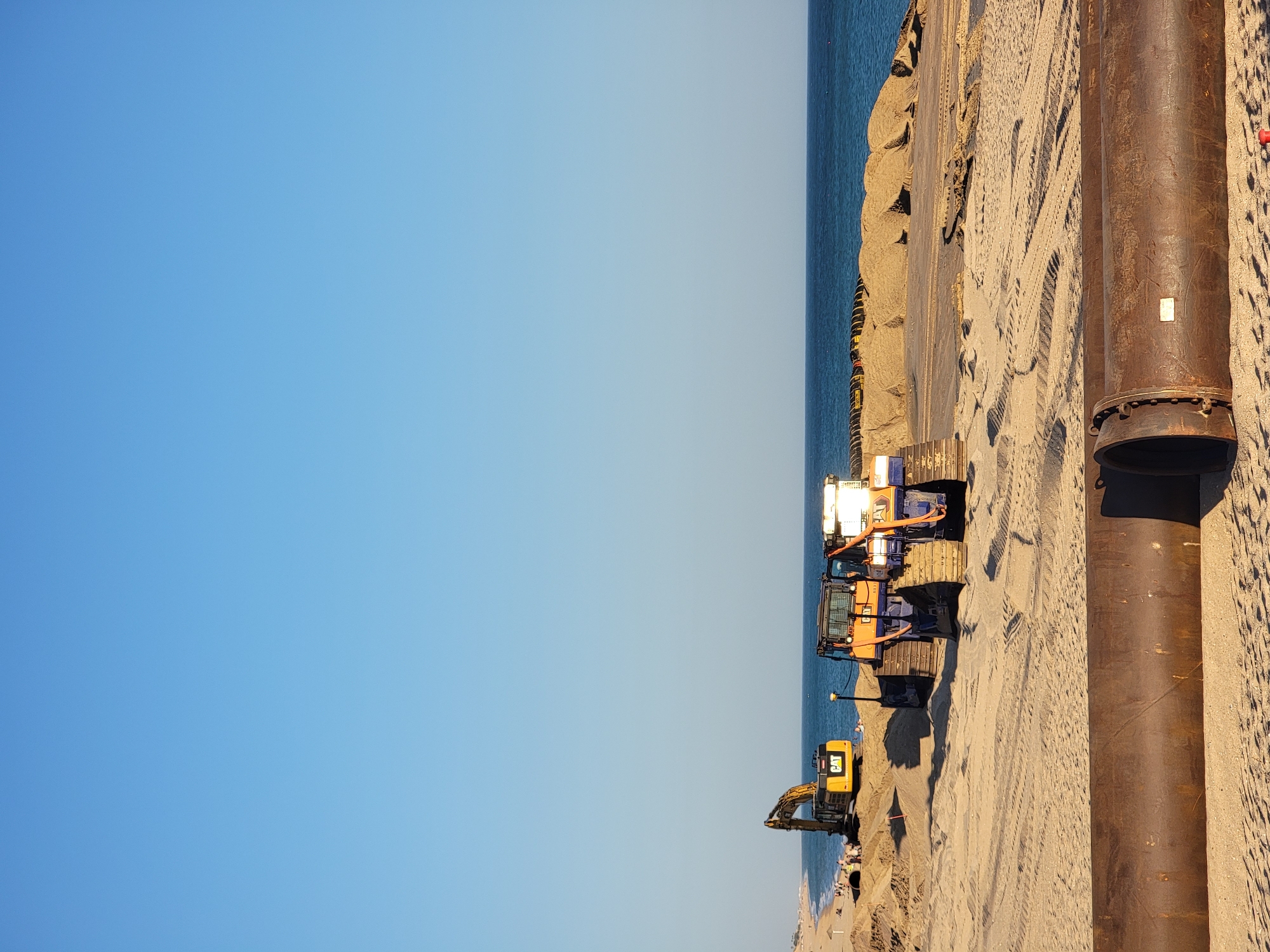 The image depicts a construction scene on a sandy beach in Quarteira, Portugal. In the foreground, two large, rusty metal pipes lie horizontally on the sand. In the midground, several construction vehicles are present, including a yellow excavator with "CAT" branding and an orange and blue tracked vehicle also bearing "CAT" branding. These vehicles are positioned amidst mounds of sand. In the background, the blue ocean stretches to the horizon under a clear, pale blue sky. A faint line of beachgoers and umbrellas is visible on the distant shoreline. The overall impression is one of ongoing coastal construction or maintenance work.